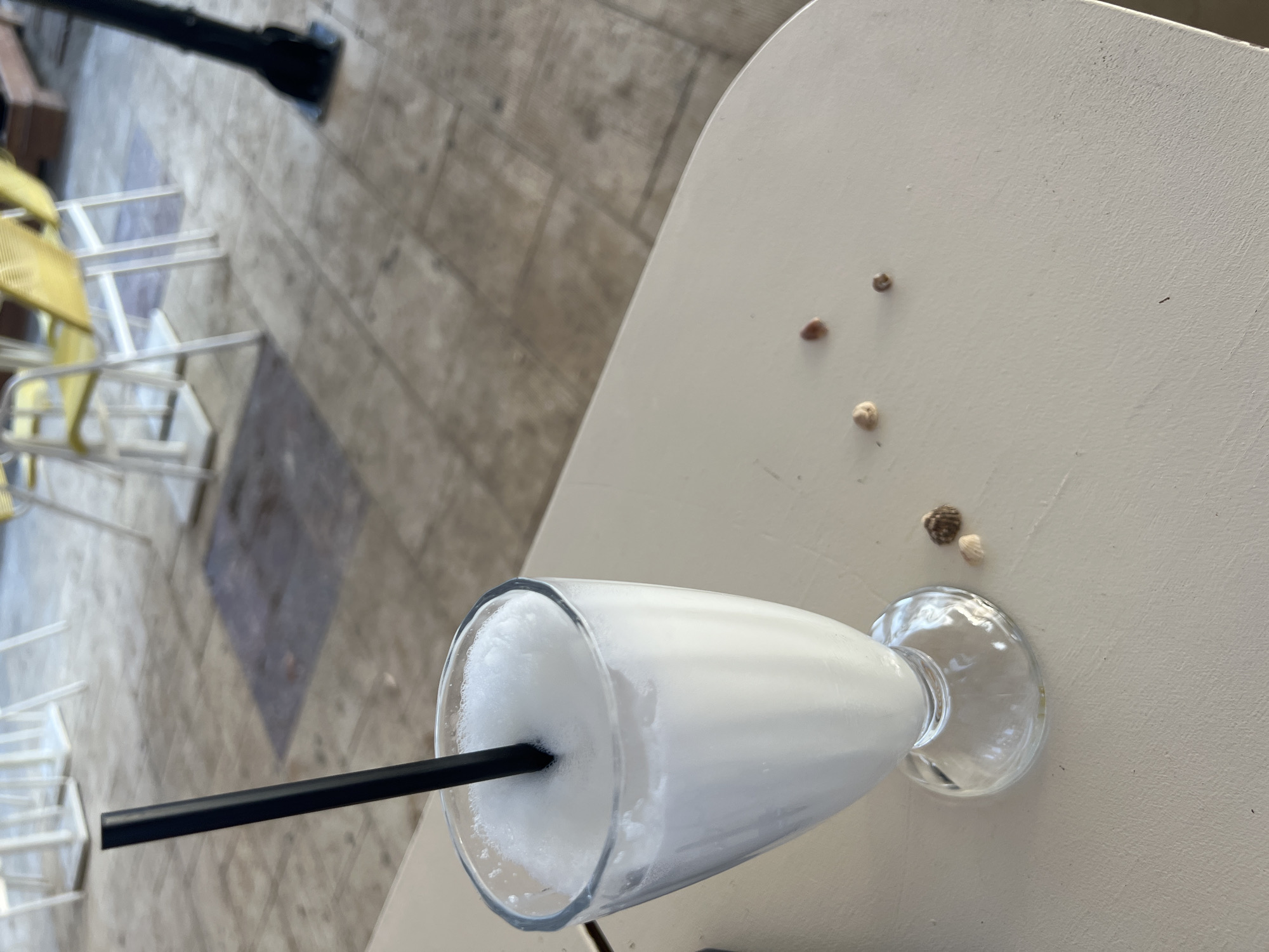 A close-up, eye-level shot shows a clear glass filled with a frothy, white beverage, likely a milkshake or smoothie, with a black straw inserted. The glass rests on a light-colored, textured table. To the right of the glass are a few small, light-colored objects that appear to be seashells or pebbles. In the background, the scene is out of focus, revealing a patio or outdoor seating area with light-colored tile flooring and several white tables and yellow chairs. The overall setting suggests a cafe or restaurant in a sunny, outdoor environment, possibly in Eretria Municipal Unit, Greece, given the presence of seashells and the characteristic outdoor dining culture. No people are visible in the frame.