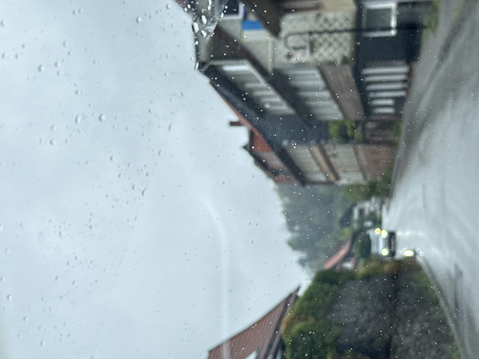 The image shows a view through a wet window, likely from inside a vehicle, of a street scene in the town of Walkenried, Germany. Raindrops are visible on the window. On the left side of the road, there is a stone wall covered with greenery, and a house with a red roof. Further down the street, a white car with its headlights on is approaching. On the right side of the road, there are several buildings with multi-story facades, typical of a European town. The sky is overcast, suggesting a rainy day. The road appears wet and reflective, consistent with rainfall. The overall impression is a quiet, overcast day in a residential or mixed-use area of Walkenried.