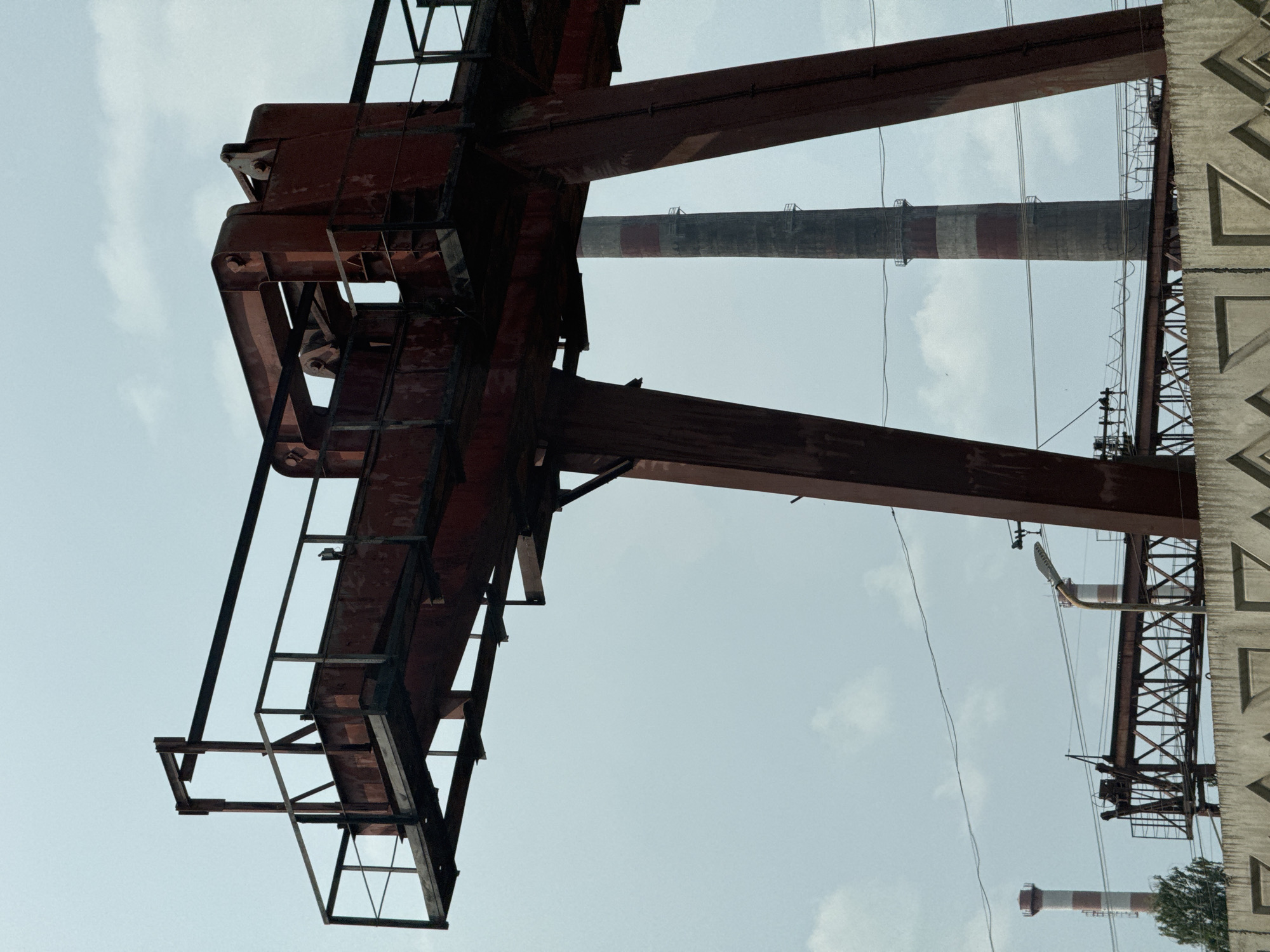This is a photograph taken from a low angle, looking upwards at an industrial setting. The primary subject is a large, rusty red metal structure, likely a gantry crane, dominating the left side of the frame. It is made of thick I-beams and appears to be a weathered, industrial piece of equipment.
In the background, two smokestacks are visible. The larger one is closer and appears to be a traditional industrial chimney with red and grey bands. The smaller one, further in the distance, also has red and white bands. Wires and support structures for power lines are also present, crisscrossing the sky.
On the far right of the image, a portion of a concrete structure with a geometric, repeating pattern is visible. This could be part of a building or an industrial wall. The sky is overcast with some patches of white clouds, suggesting a daytime scene. The overall mood is one of industrial scale and perhaps a sense of decay due to the rusted metal. No people are visible in the image. There is no visible text. Given the location context of Izhevsk, Russia, this is likely a view of an industrial zone or a factory complex.