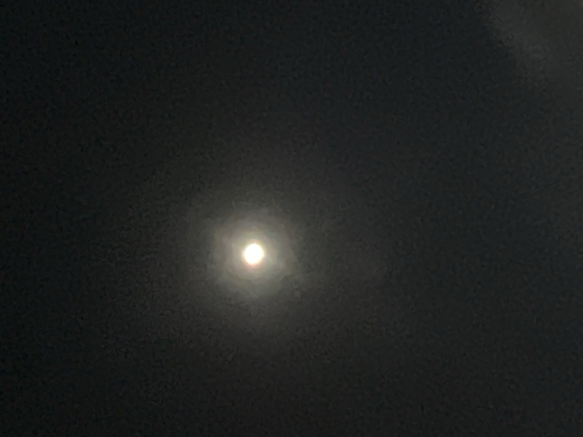 A full moon is visible in the night sky, centered in the frame. The moon is bright and appears to be surrounded by a soft glow or halo, suggesting it is partially obscured by clouds or atmospheric haze. The background is dark, characteristic of a nighttime scene. The overall image quality is grainy, likely due to low light conditions and the resolution of the camera. There is no discernible indication of the city of Denpasar, Indonesia, or any specific environmental or human elements within the frame.