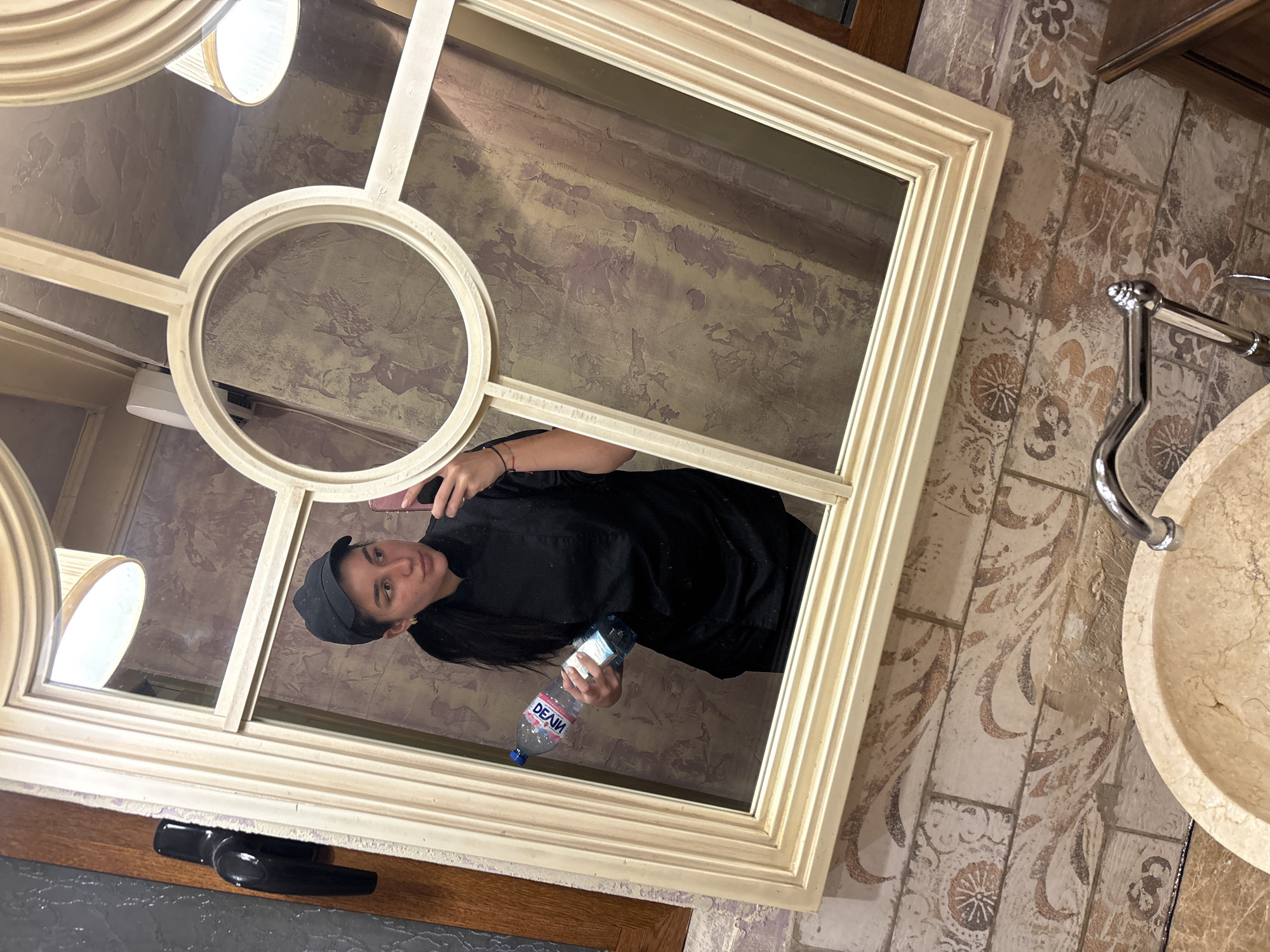 A reflection in a rectangular mirror framed in cream-colored molding shows a person wearing a black outfit and a gray cap, holding a water bottle labeled "DEVIN" in their right hand and a smartphone in their left. The mirror is mounted on a wall with textured, cream-colored plaster, decorated with purple and brown swirls. To the right of the mirror, patterned tiles in shades of brown and cream are visible, along with a portion of a marble sink and chrome faucet. The lower portion of the mirror frame is mounted on a dark wood surface. The location may be Veliko Tarnovo, Bulgaria.
