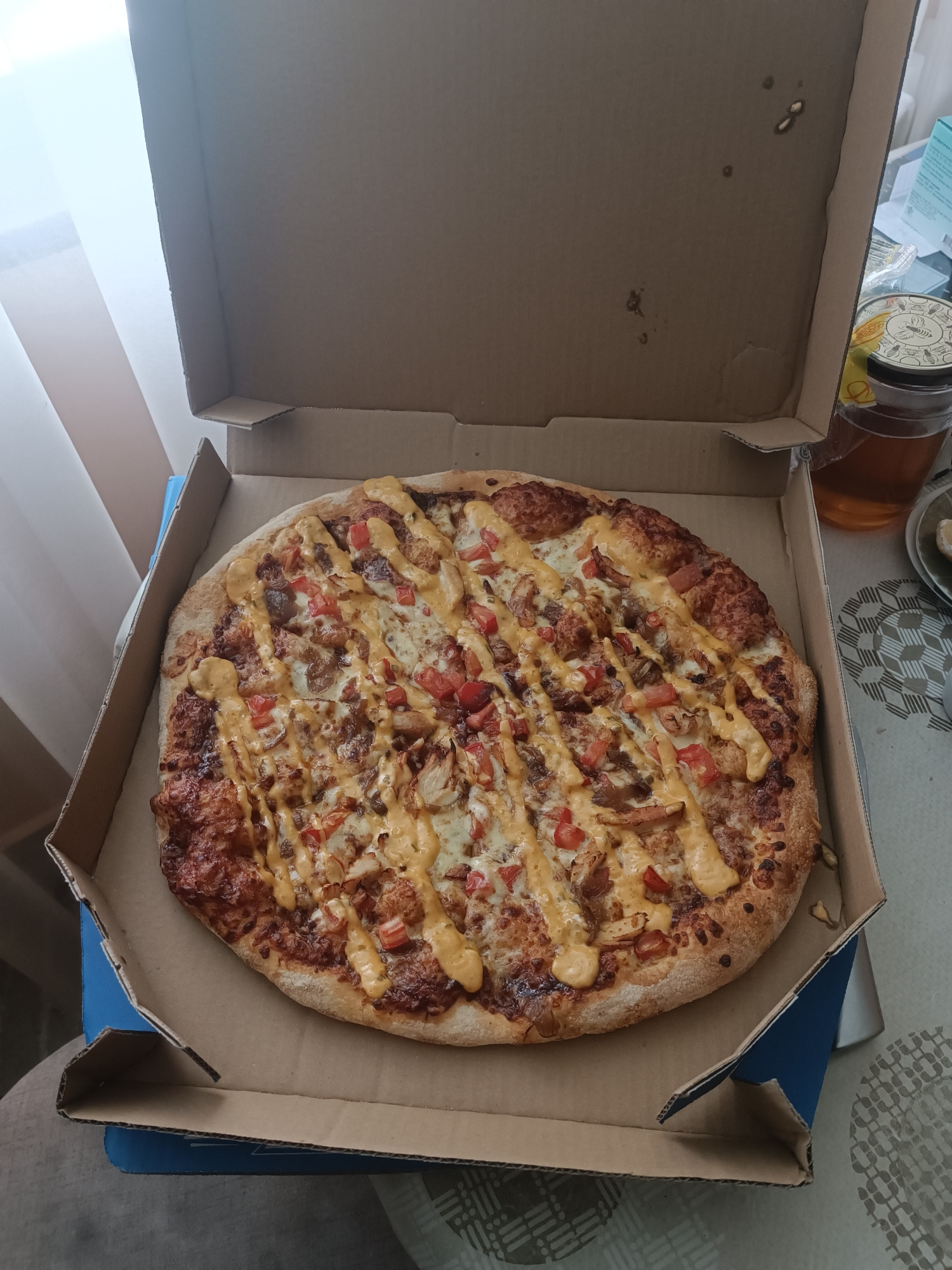 A close-up, eye-level shot shows a freshly baked pizza in an open cardboard pizza box. The pizza is round and appears to be topped with cheese, red diced tomatoes, pieces of meat, and a drizzle of a creamy orange sauce. The crust is golden brown and slightly puffed. The pizza box is made of corrugated cardboard, with some blue coloring visible on the sides of the bottom portion. The background is blurred, but suggests an indoor setting with natural light coming from a window on the left. To the right of the pizza box, a jar of honey with a decorative lid and a glass of amber liquid are partially visible. The table surface is covered with a patterned tablecloth. The scene is consistent with a meal being served at a residence in Burgas, Bulgaria, on a daytime.