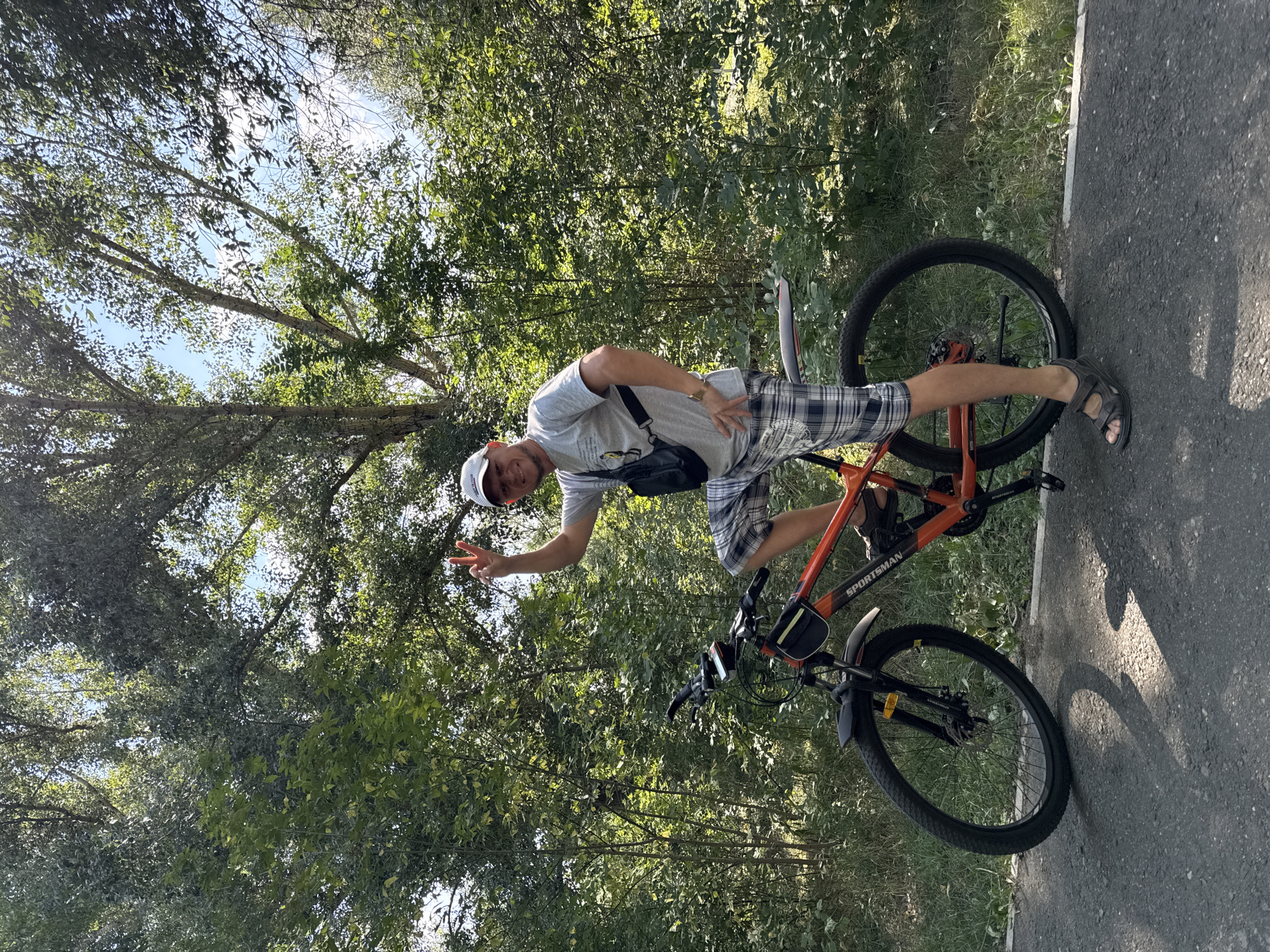 The image shows a man on a bicycle in a park. The man is wearing a grey t-shirt, plaid shorts, a white baseball cap, and dark sandals. He is holding up two fingers on his right hand in a peace sign and smiling. The bicycle is orange and black, and it has a black bag attached to the frame. The man is standing on the bicycle pedals with his left foot. The background of the image is a lush green forest with trees and foliage. The ground is a paved path. There is a shadow cast on the path from the bicycle. The image appears to have been taken on a sunny day. The location is identified as the city of Oskemen, Kazakhstan.