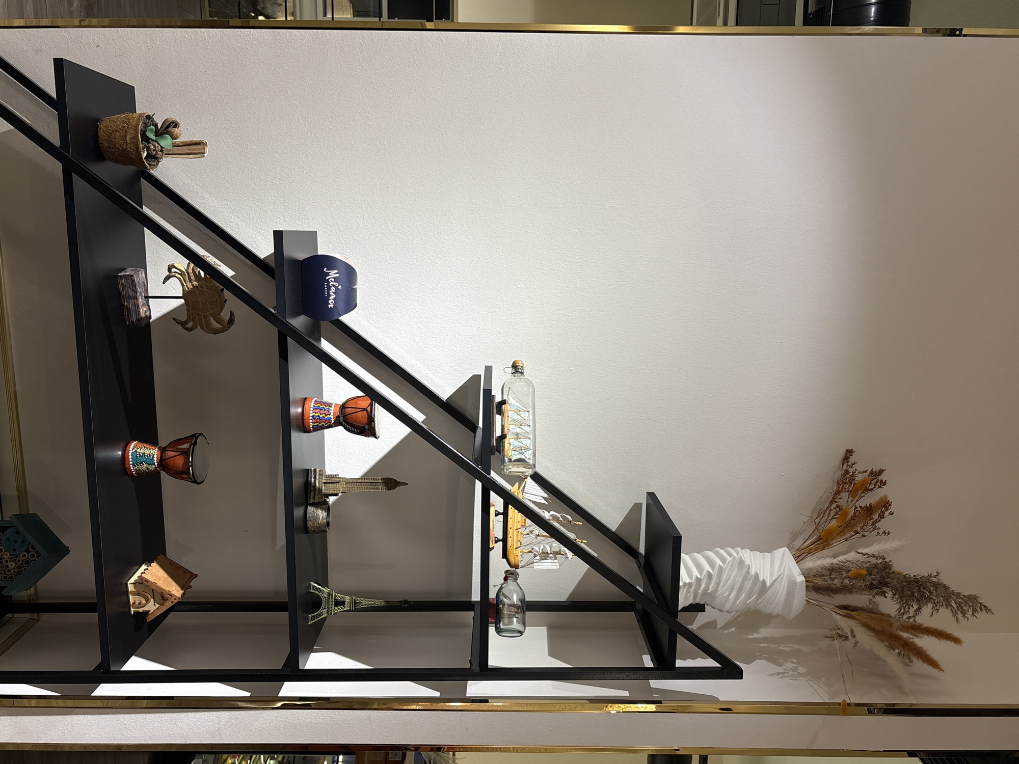 A black metal shelving unit with a stepped design against a white wall. The shelving unit is decorated with various objects. On the top shelf, a white textured vase holds a dried floral arrangement. Below that, a glass bottle contains a model ship, and another smaller glass bottle holds a miniature scene. The third shelf displays a small Eiffel Tower replica, a miniature skyscraper, and a small metal cup. The fourth shelf holds a decorative crab sculpture, a small potted plant, and a blue box with white text. The bottom shelf features two decorative drums and a miniature wooden house. A narrow gold trim runs vertically along the right edge of the shelving unit. The lighting suggests an indoor setting, possibly a retail store or home. The scene does not contain any people or actions.