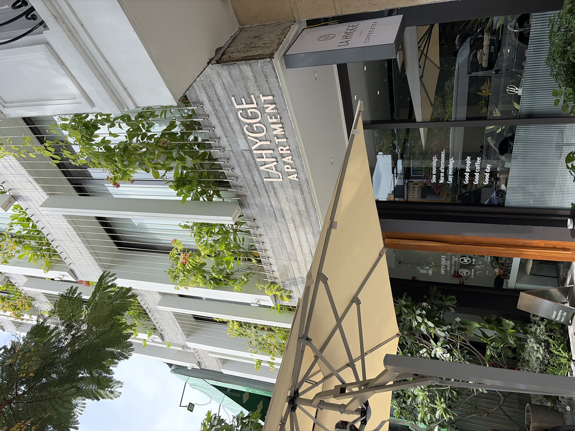 This image shows the exterior of a building with the signage "LAHYGGE APARTMENT" and "LA HYGGE COFFEE & TEA" visible. The building features a modern architectural design with white vertical elements and lush green plants cascading down the facade. An outdoor umbrella is partially visible in the foreground, suggesting a cafe or seating area. The reflections in the glass windows of the building show a street scene with vehicles and other buildings, indicative of an urban environment. Based on the signage and architectural style, this location is likely in Đà Nẵng, Vietnam. There are no people visible in the image.