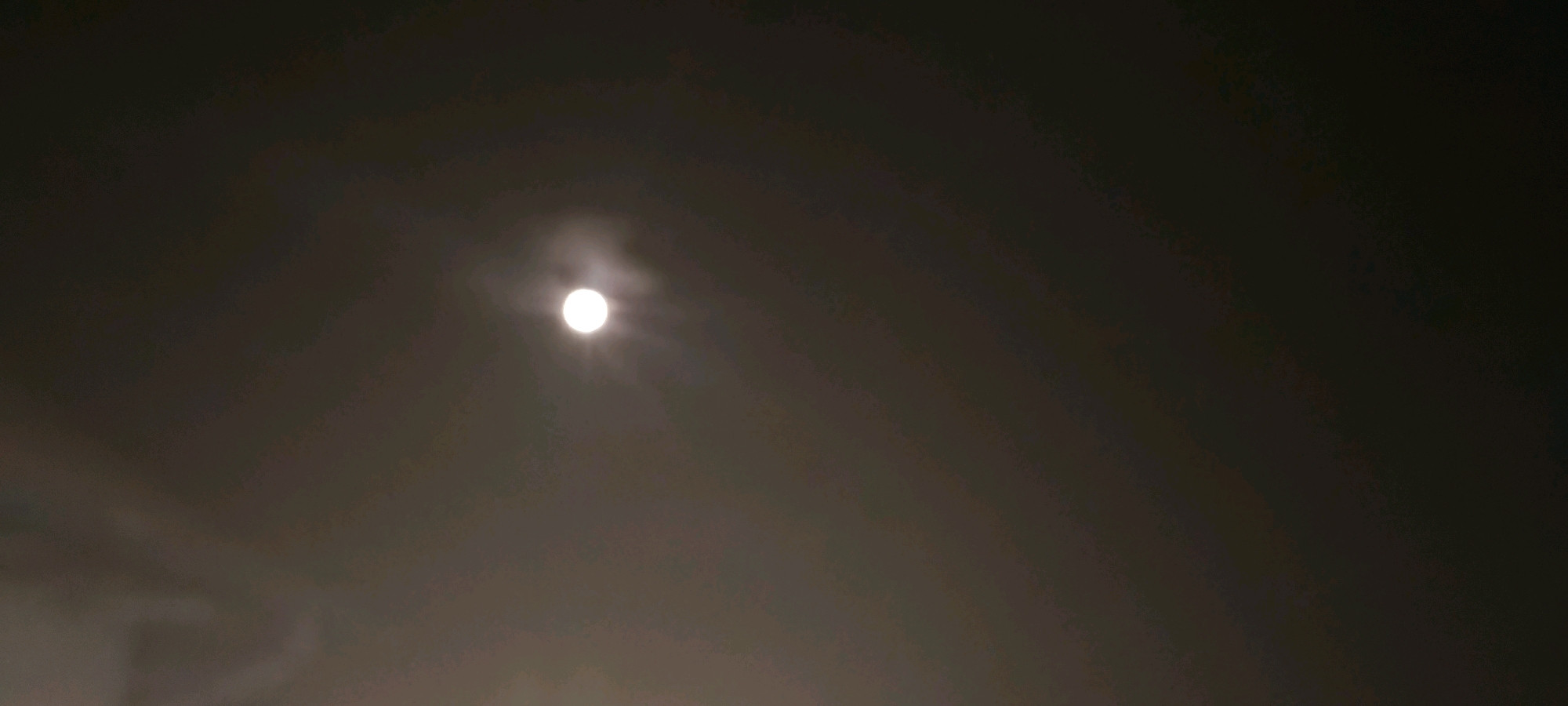 The image is a photograph of a full moon against a dark, cloudy sky. The moon is bright and luminous, with a soft halo of light surrounding it. Wisps of clouds partially obscure the moon, creating a hazy effect. The sky is a gradient of dark grays and blacks, suggesting nighttime. The image appears to be taken from ground level, looking upwards. There are no people, buildings, or specific landmarks visible in the frame. The photograph's focus is solely on the celestial body and the atmospheric conditions. There is no indication of the location in Dolni Dabnik, Bulgaria.
