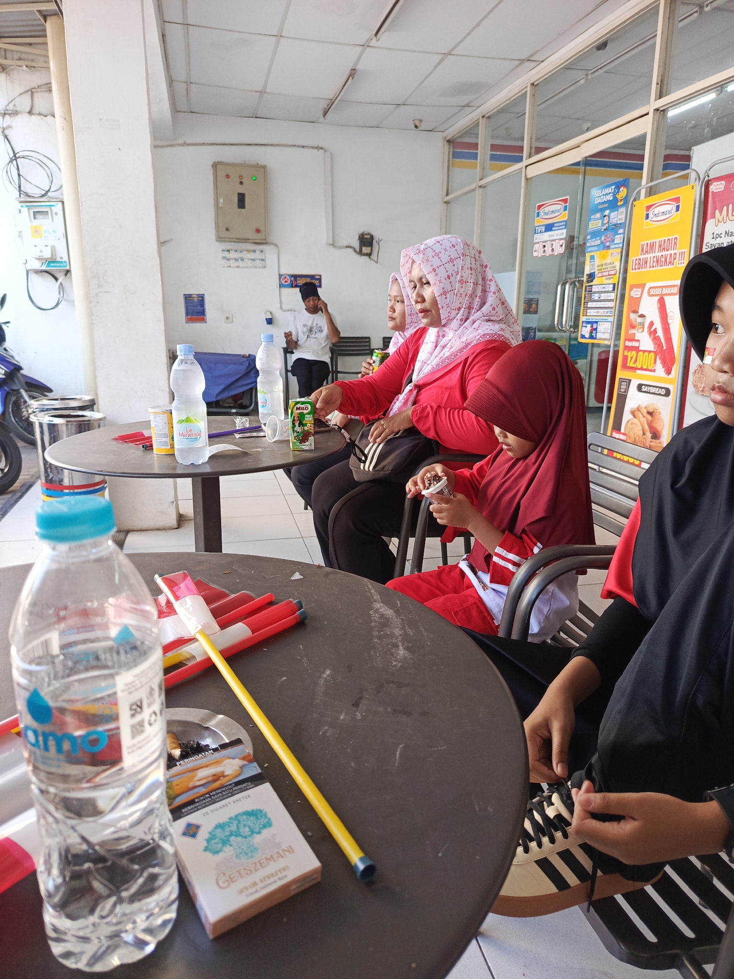 The image captures a scene at an outdoor seating area adjacent to an Indomaret convenience store in Semampir, Indonesia. In the foreground, a partially visible plastic water bottle with a blue cap sits on a dark, round table. Also on the table are a pack of GETSEMANI kretek cigarettes and an ashtray. Several red and yellow markers or sticks are also on the table. In the mid-ground, a young girl wearing a maroon hijab and matching outfit is seated, holding a small ice cream cone. Beside her, a woman in a pink hijab and red shirt is seated, looking to the left. Another woman in a maroon hijab is partially visible behind them. In the background, another Indomaret store is visible through a glass storefront, displaying promotional posters. A man in a white t-shirt is seated further back, looking at his phone. The environment suggests a casual gathering outside the store on a bright day.