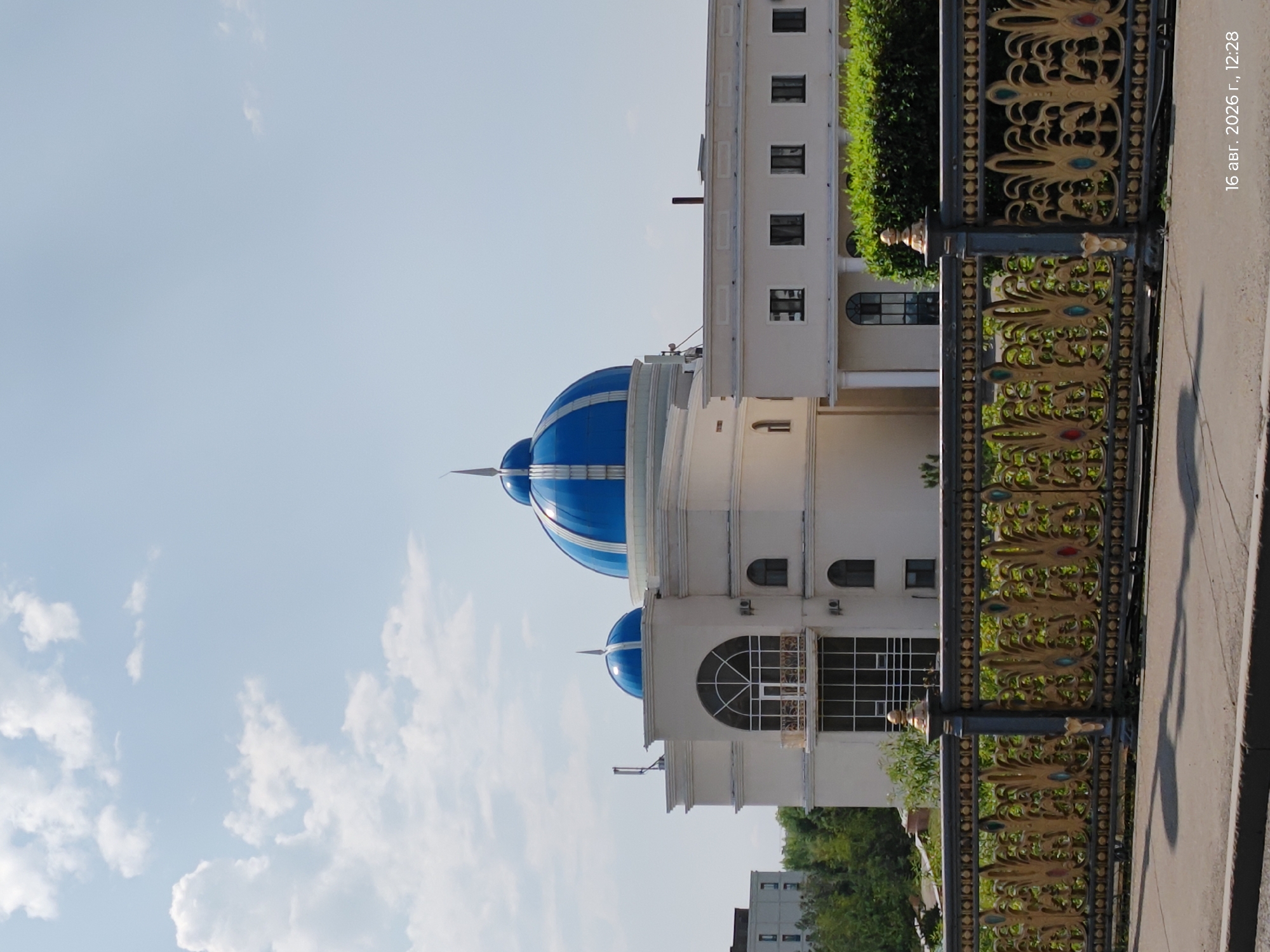 The image shows a building with blue domes in Astana, Kazakhstan. The building has a light-colored facade and multiple windows. In the foreground, there is a decorative metal fence with golden ornamentation. The sky is partly cloudy. The bottom right corner of the image displays a timestamp: "16 авг. 2026 г., 12:28". There are no people or animals visible in the image.