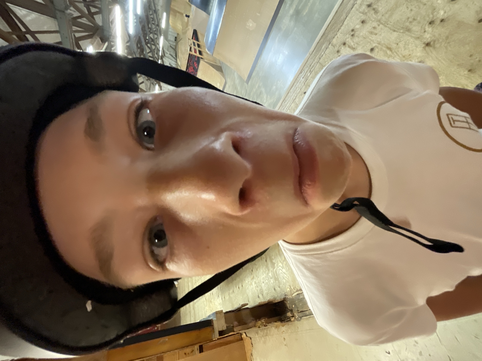 The image is a close-up, fisheye lens portrait of a young person wearing a black helmet and a white t-shirt. The subject's face is distorted by the wide-angle lens, with their eyes wide open and their mouth in a pouting expression. The background appears to be a skatepark, with wooden ramps and structures visible. The lighting suggests an indoor environment. The location is identified as Bluewater, Canada, but no specific features within the image confirm this.