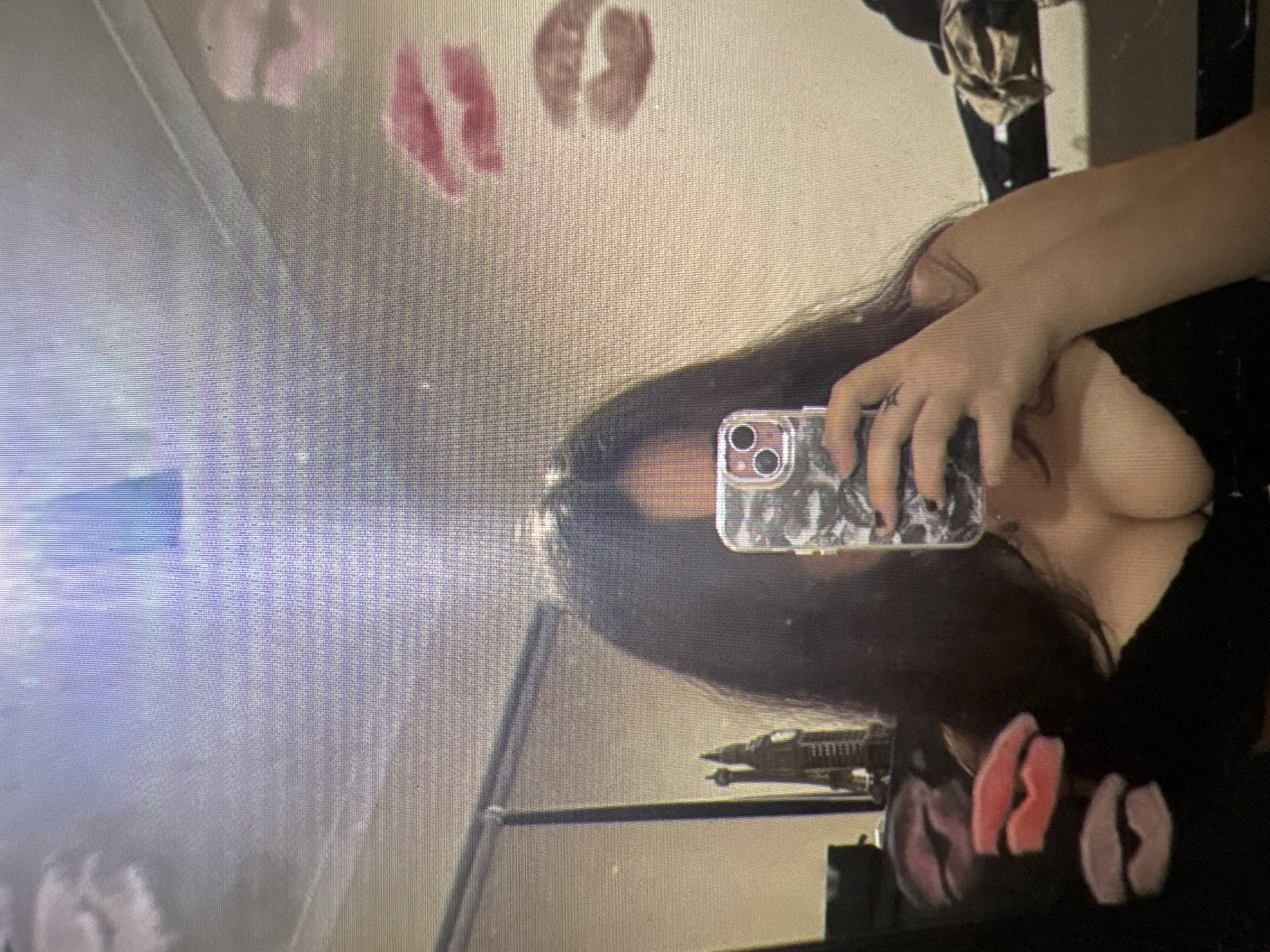 The image depicts a person taking a selfie in a room. The person has dark hair and is wearing a black top with a low neckline. They are holding a phone with a patterned case. The background shows a wall with several lipstick kiss marks. There are also objects visible, including what appears to be a miniature Eiffel Tower on the left side of the image and some clothing items on the right. The overall lighting suggests an indoor setting. The image's characteristics do not provide any specific information about the city of Castelo Branco, Portugal.