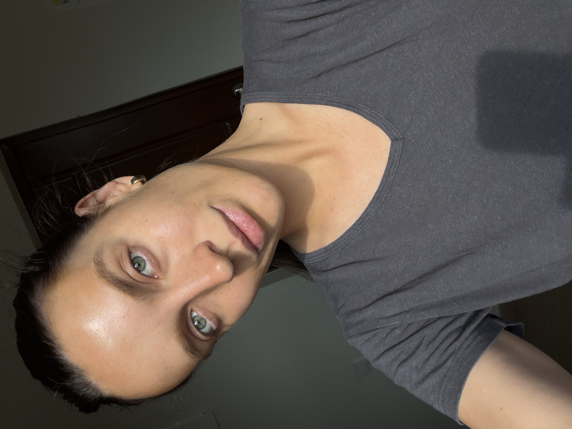 A head and shoulder shot features an individual with light skin, dark hair pulled back, and striking greenish-blue eyes, looking directly at the viewer. The person is wearing a dark grey t-shirt or casual top. A small, golden hoop earring is visible in their left ear. The individual's expression is neutral and direct, with a slight upward tilt to their head. The skin appears clear, with a natural sheen on the forehead and nose, and the lips are a soft pink. The background is dark and out of focus, suggesting an indoor setting, possibly against a dark wall or piece of furniture. A dark, wooden door or cabinet top is faintly visible behind the head and ear. The lighting highlights the face, casting subtle shadows that define the contours of the neck and collarbone. The image is oriented sideways, with the person's head rotated 90 degrees counter-clockwise from a typical upright portrait. There is no discernible activity other than posing for the photograph. No visible text is present. The location context provided is Budva, Montenegro, though there are no visual cues within the image to confirm this specific location.