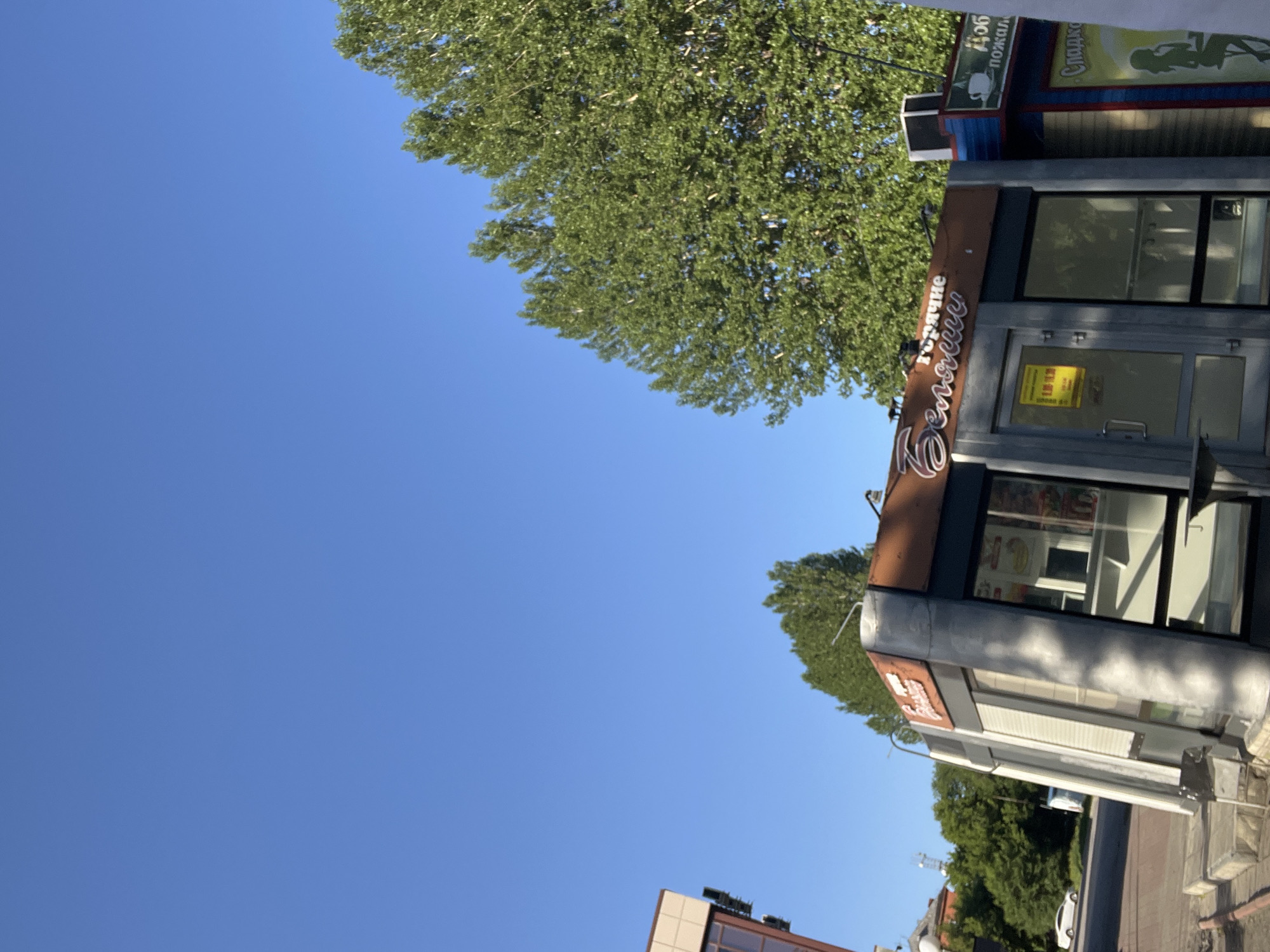 The image shows a clear blue sky over a street scene in Kiselyovsk, Russia. In the foreground, there are two kiosks. The kiosk on the left has a sign with Russian text that appears to say "Горячие" (Hot) and "Беляши" (Belyashi), which are traditional Russian pastries. The kiosk on the right has a sign with Russian text that says "доб" (short for "добро пожаловать" - welcome) and "Сладк" (short for "сладкое" - sweets). There are trees with green foliage visible behind the kiosks. A white car is parked on the street to the left of the kiosks, and a building with a beige facade is partially visible in the far left background. The scene appears to be set during daytime, with sunlight casting shadows.