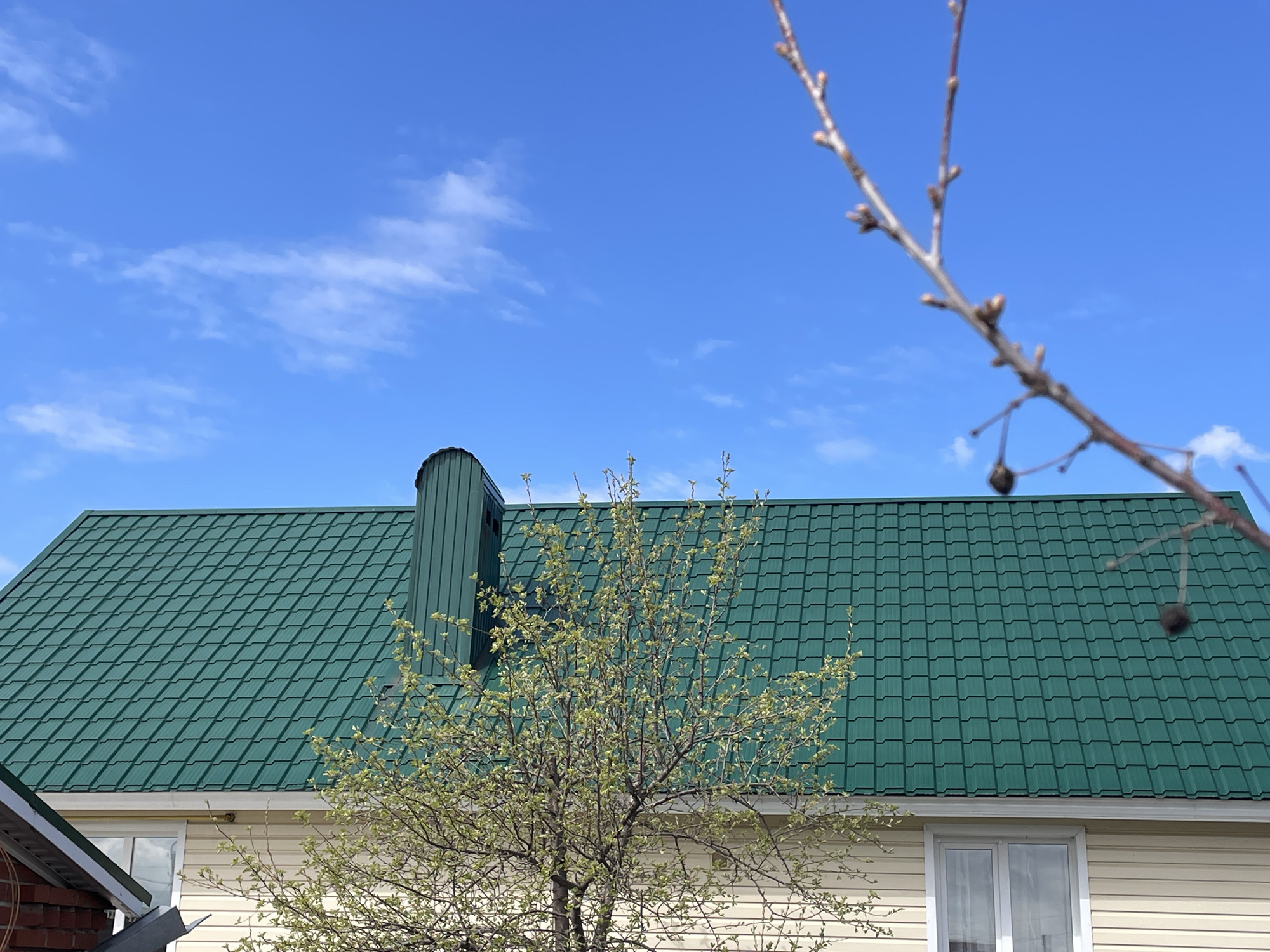 The image shows a section of a house in Oktyabrsky, Russia. The house has a green metal tile roof and beige siding. A tree with emerging spring foliage is in front of the house, partially obscuring the view. A metal chimney extends from the roof. Bare branches with buds and a few dry fruits are visible in the upper right foreground. The sky is blue with scattered white clouds. A small portion of an adjacent building is visible on the left edge. A window is visible on the side of the house.