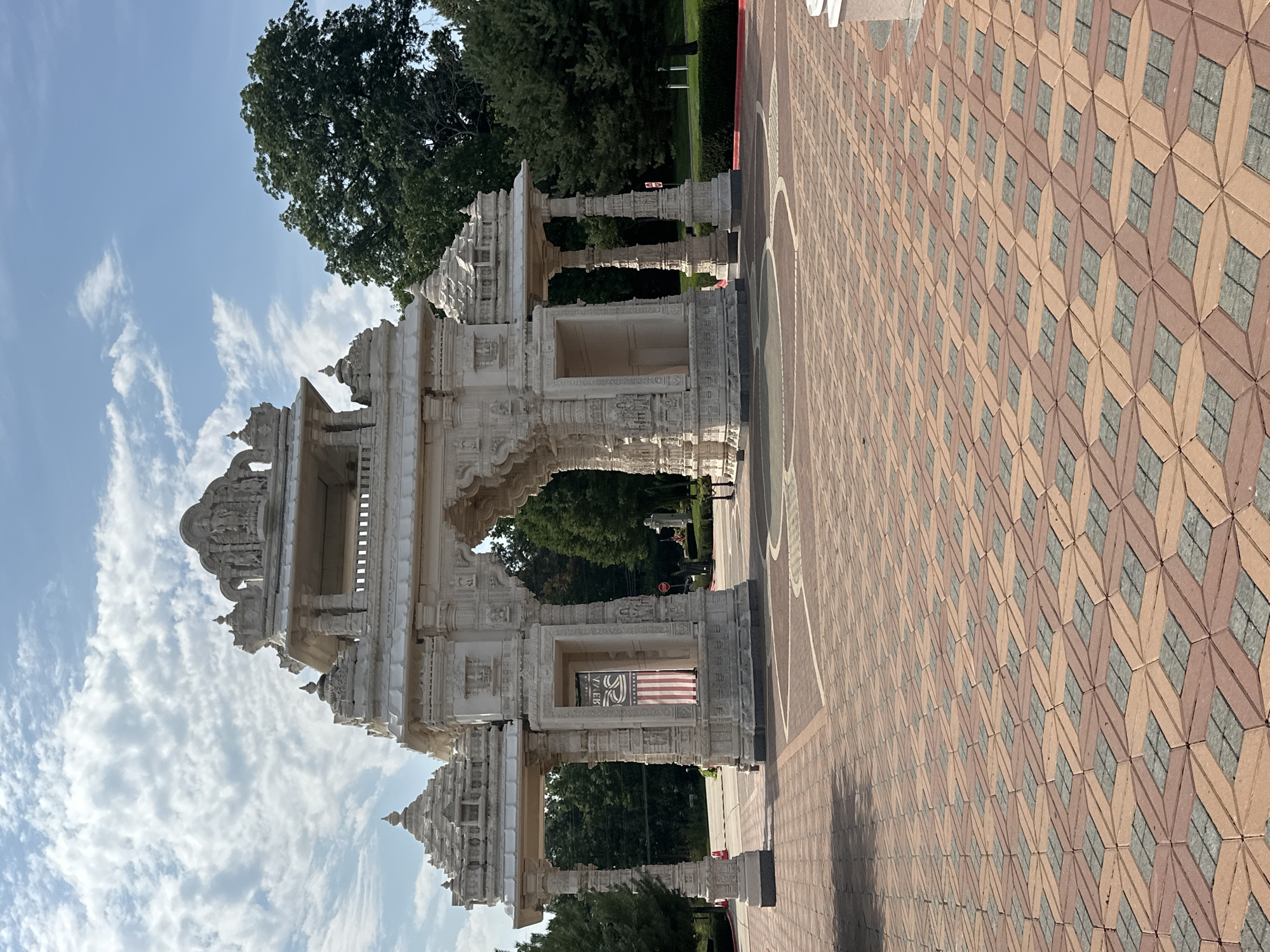 The image shows the ornate entrance archway of a Hindu temple, likely the BAPS Shri Swaminarayan Mandir in Bartlett, Illinois, United States of America. The archway is constructed of white stone and features intricate carvings and tiered roofs characteristic of traditional Indian architecture. An American flag is visible hanging within an alcove of the arch. The ground in front of the arch is paved with red and grey interlocking bricks arranged in a geometric pattern. Lush green trees form the backdrop, and a bright blue sky with scattered white clouds completes the scene. The overall impression is one of grandeur and cultural significance.