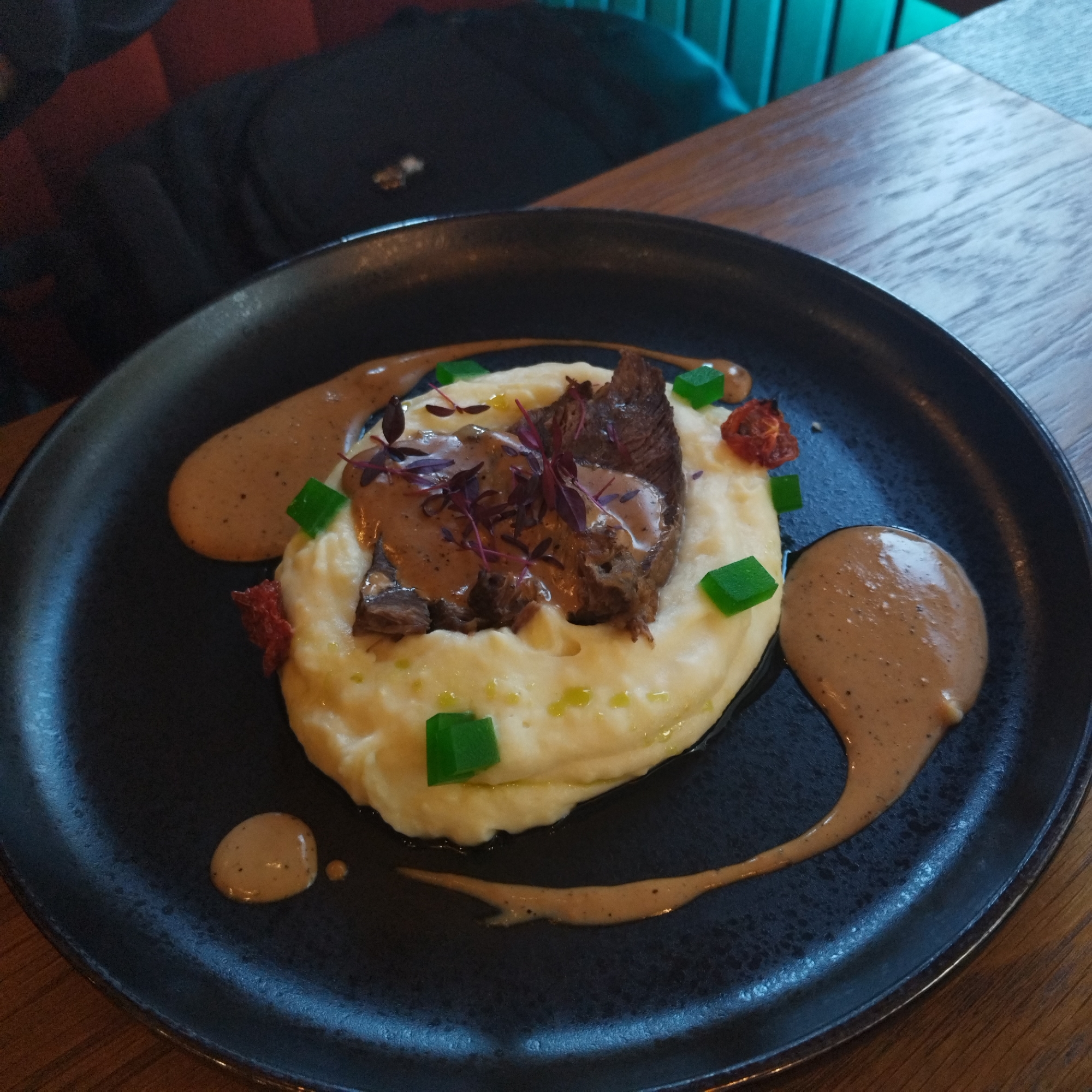 The image displays a close-up, top-down view of a meticulously presented meal on a dark, textured ceramic plate. At the center, a generous mound of creamy white mashed potatoes or a similar puree forms the base. Resting atop the puree are several pieces of dark, braised or slow-cooked meat, generously coated in a smooth, light brown gravy speckled with darker particles.
The dish is adorned with several garnishes for color and texture. Delicate purple microgreens or herbs are scattered over the meat, while small, vibrant green translucent cubes, possibly a gelée or compressed vegetable, are placed around the mashed potatoes. A few dried red pieces, resembling sun-dried tomatoes or roasted peppers, also punctuate the presentation. A speckled, light brown sauce is elegantly drizzled in swirls around the main food elements on the plate.
The background is softly blurred, revealing parts of an indoor dining setting. A warm wooden table surface is visible, alongside indistinct shapes suggesting restaurant seating, including dark fabric with hints of red and teal/green accents. This indicates the meal is being served in a restaurant or cafe, likely in Yekaterinburg, Russia, as per the context. The lighting suggests a well-lit environment, either daytime or evening. There is no visible text in the image, nor are any specific weather conditions or emotions discernible.