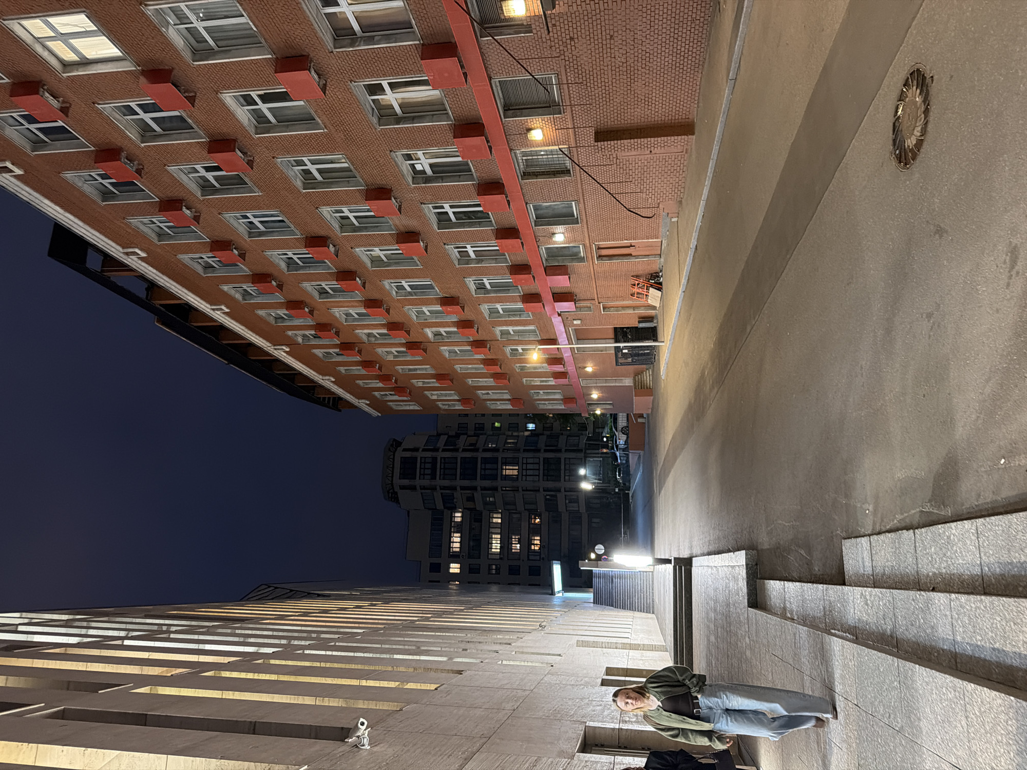 A woman walks along a tiled path next to a modern building with vertical light fixtures on its left. On the right is a brick building with numerous windows and red rectangular protrusions above some of them. In the distance, several other buildings are visible, including a tall, multi-story structure with illuminated windows. A street extends into the distance, with streetlights and parked cars. The scene is illuminated by artificial light, and the sky is dark, indicating it is nighttime. The location is in Moscow, Russia.