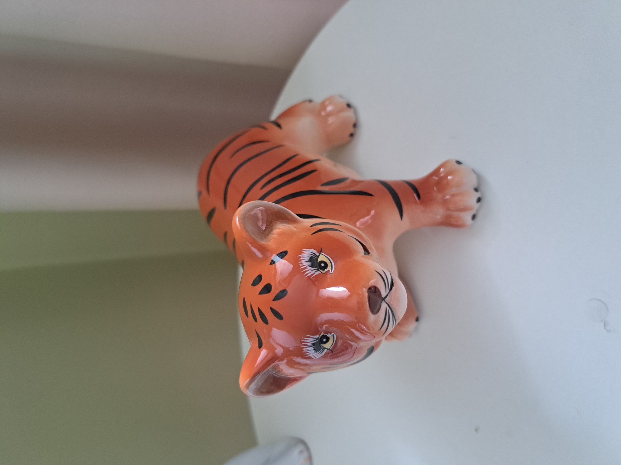 A ceramic figurine of a tiger with orange and black stripes is placed on a white surface. The tiger is in a crouching position, with its head facing forward and its eyes looking directly at the camera. The figurine has exaggerated eyelashes. The background consists of a pale green wall on the left and a beige curtain on the right. The scene is indoors and the lighting is soft. There is no indication of the location in Irkutsk, Russia.