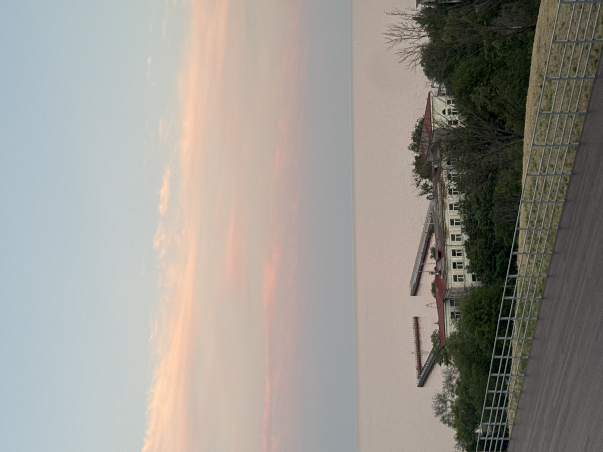 The image shows a wide, panoramic view of the sea and a coastline. In the foreground, there is a wooden walkway with a metal railing. Beyond the walkway, there are trees and buildings, with a complex of structures extending into the water. The sky is a pale blue with soft pink clouds, suggesting either sunrise or sunset. The scene appears to be a waterfront area, possibly a park or a recreational space, located in the city of Mariupol, Ukraine. The buildings and piers suggest a harbor or a coastal settlement. The overall atmosphere is serene and tranquil.