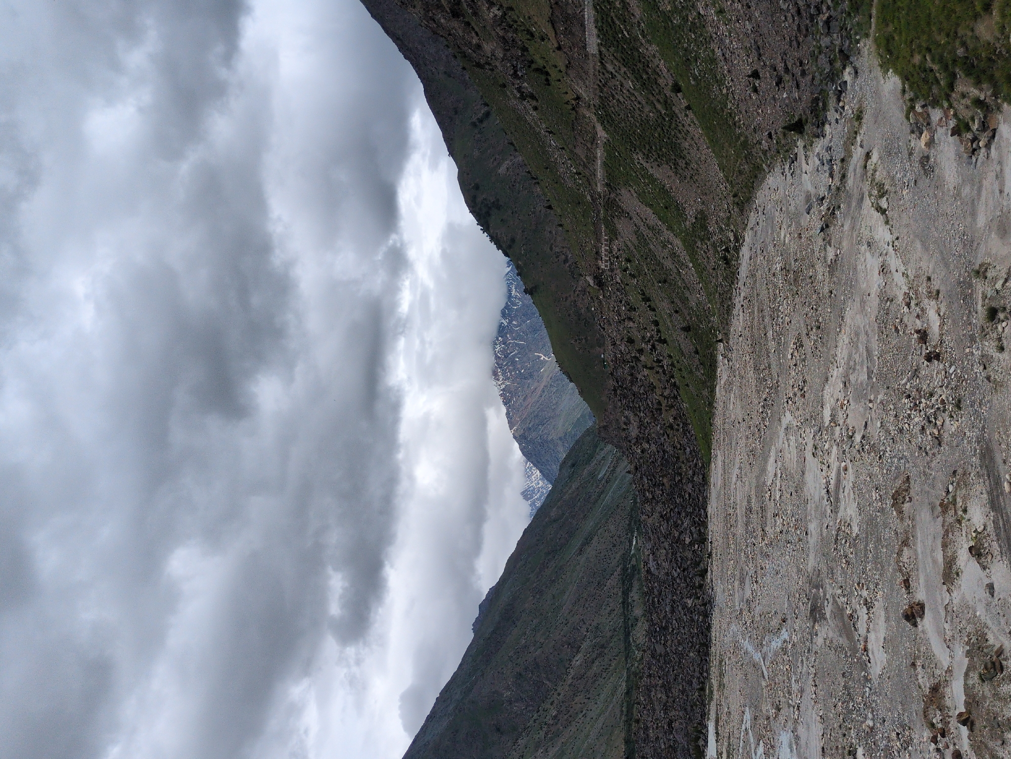 The media file depicts a dramatic mountain landscape in Darcha, India, under a cloudy sky. The primary subjects are the imposing, steep mountainsides and a dry riverbed or scree slope. The scene is characterized by rugged terrain, with the mountains covered in sparse vegetation and rocky outcrops.
The right side of the image features a wide, light-colored scree slope, heavily textured with rocks and pebbles of various sizes. Streaks of lighter and darker sediment suggest past water flow. The left side of the image is dominated by a dark, steep mountain face, also covered in rocks and patches of green vegetation. Further back, a glimpse of another mountain peak is visible, partially obscured by clouds, with snow-capped sections indicating high altitude.
The weather appears overcast, with thick, grey clouds filling the upper portion of the frame, creating a subdued and somewhat dramatic atmosphere. The lighting suggests it is daytime, but the clouds prevent direct sunlight. The overall mood is one of raw, natural beauty and the immense scale of the mountainous environment.
There are no visible people or specific objects that indicate a particular activity or event, other than the natural geological processes at play. The location cues, such as the mountain ranges and the general aridness of the scree slope, are consistent with a high-altitude, mountainous region like Darcha. No text is visible in the image.