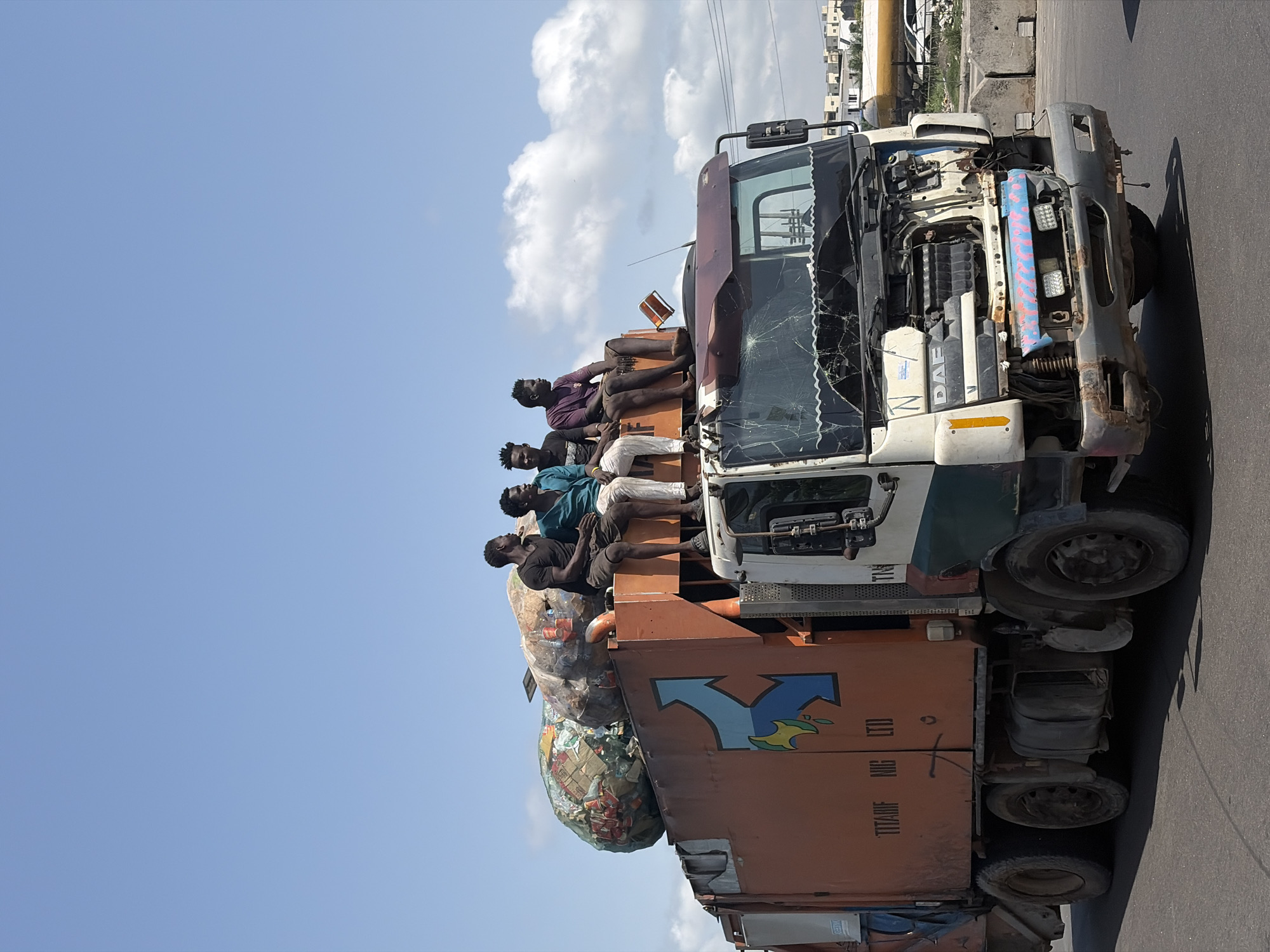 The image captures a group of men riding atop a heavily utilized truck on a sunny day in what appears to be a semi-urban or rural environment in Alaguntan, Nigeria.
**Subjects:** At least five men are visible, seated on the roof of the truck's cab and the front part of its cargo bed. They are dressed in casual attire, including shirts and shorts. The primary object is a large, older model truck, identified by the "DAF" logo on its front. The truck's front windshield is extensively cracked, and parts of its front bumper and grill are missing, exposing internal engine components. Piled on the forward section of the cargo bed are several large, bulging bags, possibly containing recyclables or other bulk materials. In the background, residential or commercial buildings can be seen under a partly cloudy sky, along with overhead power lines.
**Activity or event taking place:** The men are engaged in informal transportation, riding on the exterior of the truck, possibly traveling to a destination or being transported as part of a work crew. Their postures suggest they are either resting or simply enduring the journey. The truck appears to be on a paved road, implying active travel or a temporary stop.
**Notable details:** The scene is illuminated by bright daylight, with clear shadows indicating midday sun. The sky is predominantly blue with scattered white clouds. The truck exhibits significant wear and tear, including rust, faded paint, and structural damage to its front. The damaged windshield is particularly prominent. The large bags on the cargo bed suggest the truck is hauling cargo, and the men might be associated with this cargo or the truck's operation.
**Visible text:** The brand name "DAF" is visible on the front of the truck's cab. On the side of the truck's orange cargo bed, the faded text "TITANF NIG LTD" is discernible.