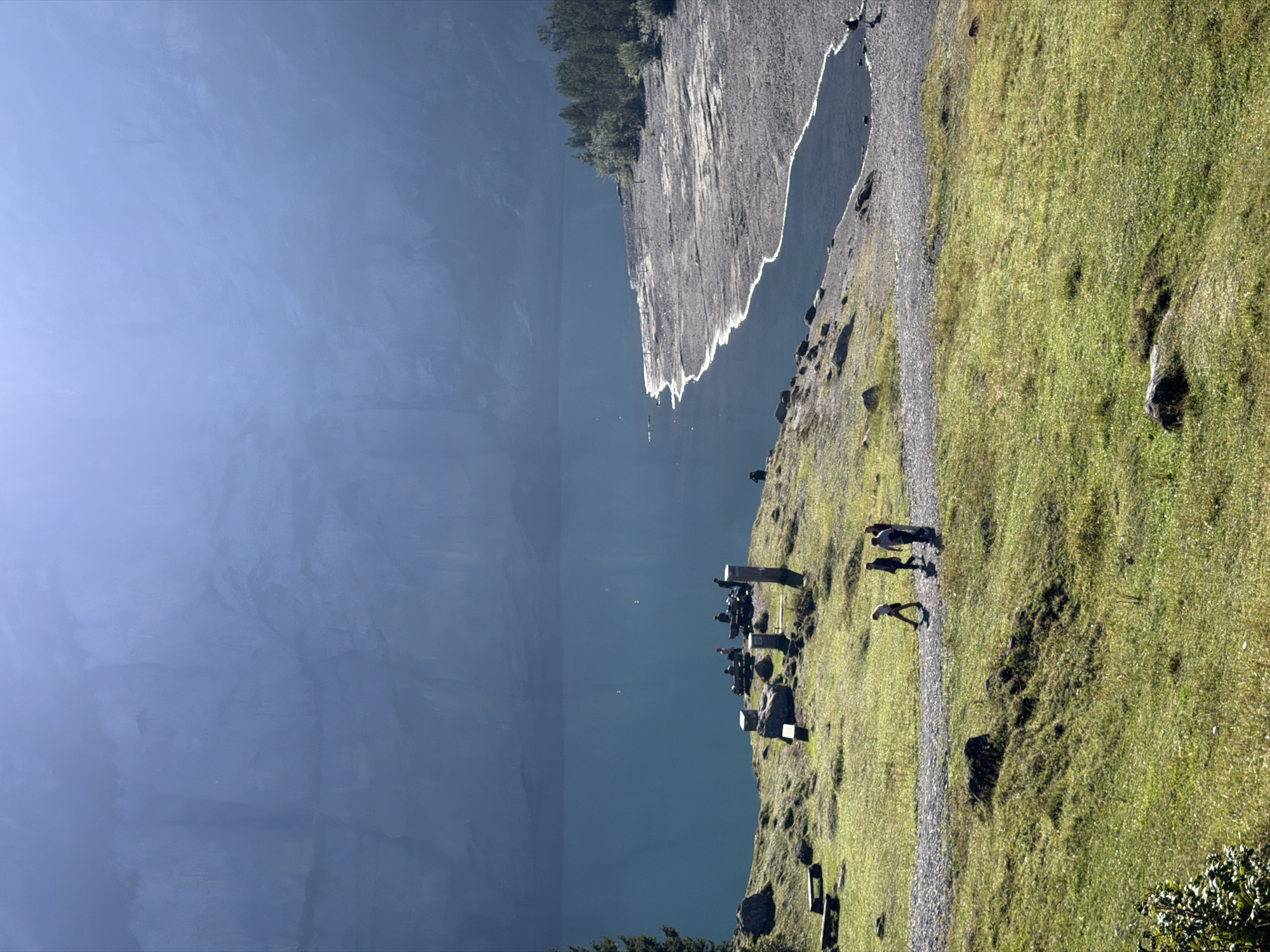 This image depicts a scenic view of a lake and surrounding landscape, likely Oeschinensee, near Kandersteg, Switzerland. The foreground features a grassy slope with scattered rocks and a pathway. Several people are walking along this path, appearing to be tourists enjoying the scenery.
Further back, there's a section of the path with several people gathered, some sitting on benches and others standing near what appear to be memorial stones or markers. These are positioned on a grassy outcrop overlooking the lake.
The lake itself is a striking, deep blue-green color, bordered by a rocky, partially exposed shoreline in the mid-ground. Beyond the shoreline, a dense forest of evergreen trees lines the edge of the lake. The background is dominated by a large, imposing mountain face, shrouded in a soft, atmospheric haze that gives it a majestic, almost ethereal quality. The lighting suggests it is daytime, with sunlight casting shadows and highlighting the textures of the landscape.