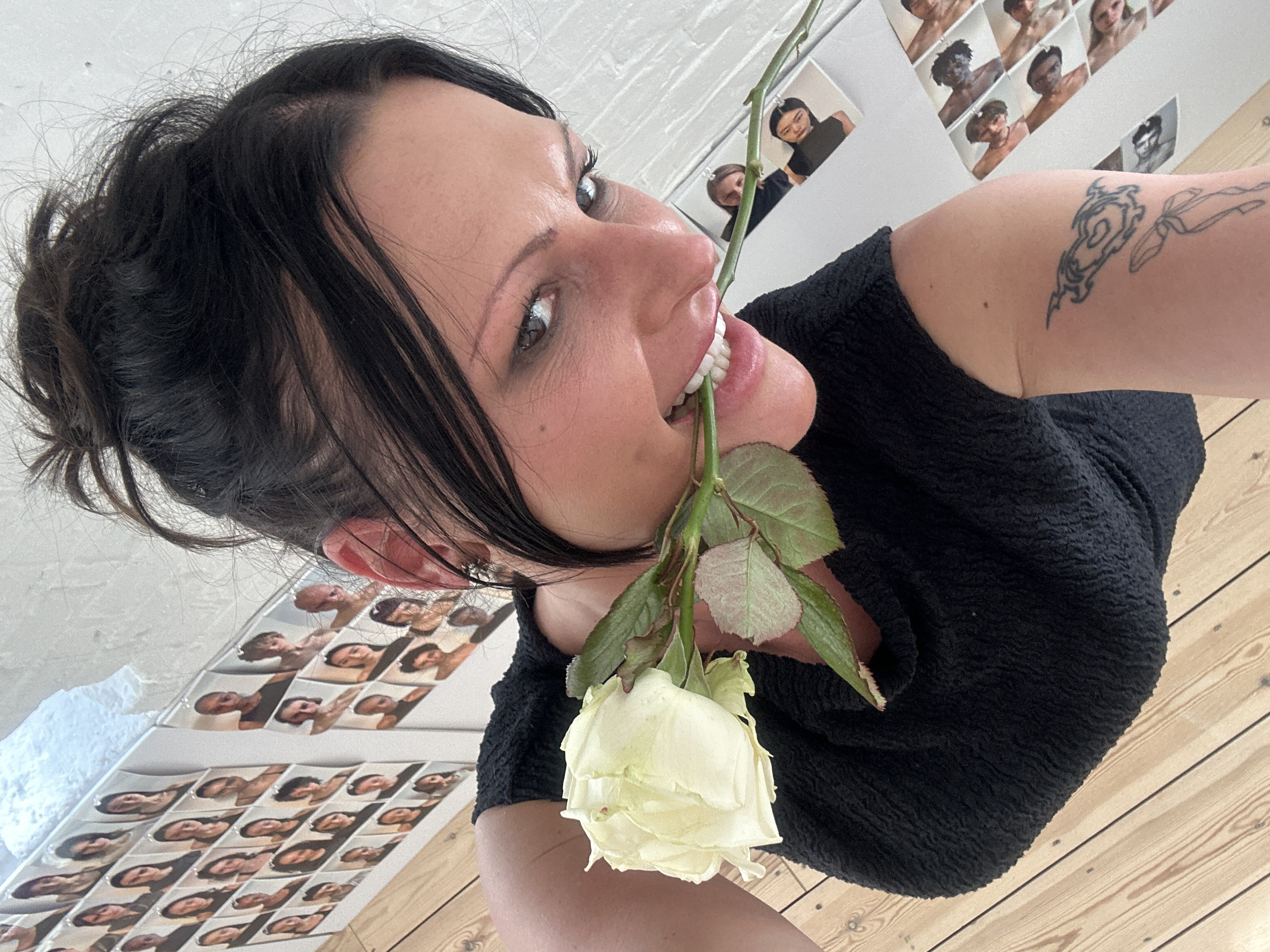 The image depicts an indoor scene focused on an adult female from a low-angle perspective. The female has dark hair, styled in an updo with loose strands, and prominent dark eye makeup. A small mole is visible below her right eye, and her right ear features a spike-style earring. She is wearing a black, textured, sleeveless top. A pale yellow rose, including its green stem and leaves, is held horizontally in her mouth, gripped by her teeth. Two tattoos are present on her left upper arm: an abstract, tribal-like design and a simpler line drawing underneath. The background comprises a light-colored wooden floor with visible planks and nail heads, leading up to a white wall. This wall is densely covered with numerous small, unframed photographic prints, predominantly headshots and upper torsos of various individuals. The wall surface behind the prints appears to have a subtle white brick texture in some sections. No specific architectural or environmental features identifying the location as London, United Kingdom, are discernible within the frame.