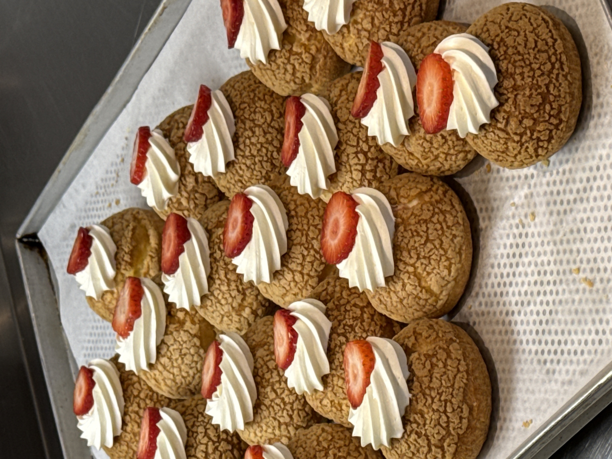 The image shows a tray of cream puffs topped with whipped cream and a slice of strawberry. The cream puffs have a cracked, golden-brown surface. The arrangement is on a metal tray lined with white parchment paper. The location of the image is unknown.
