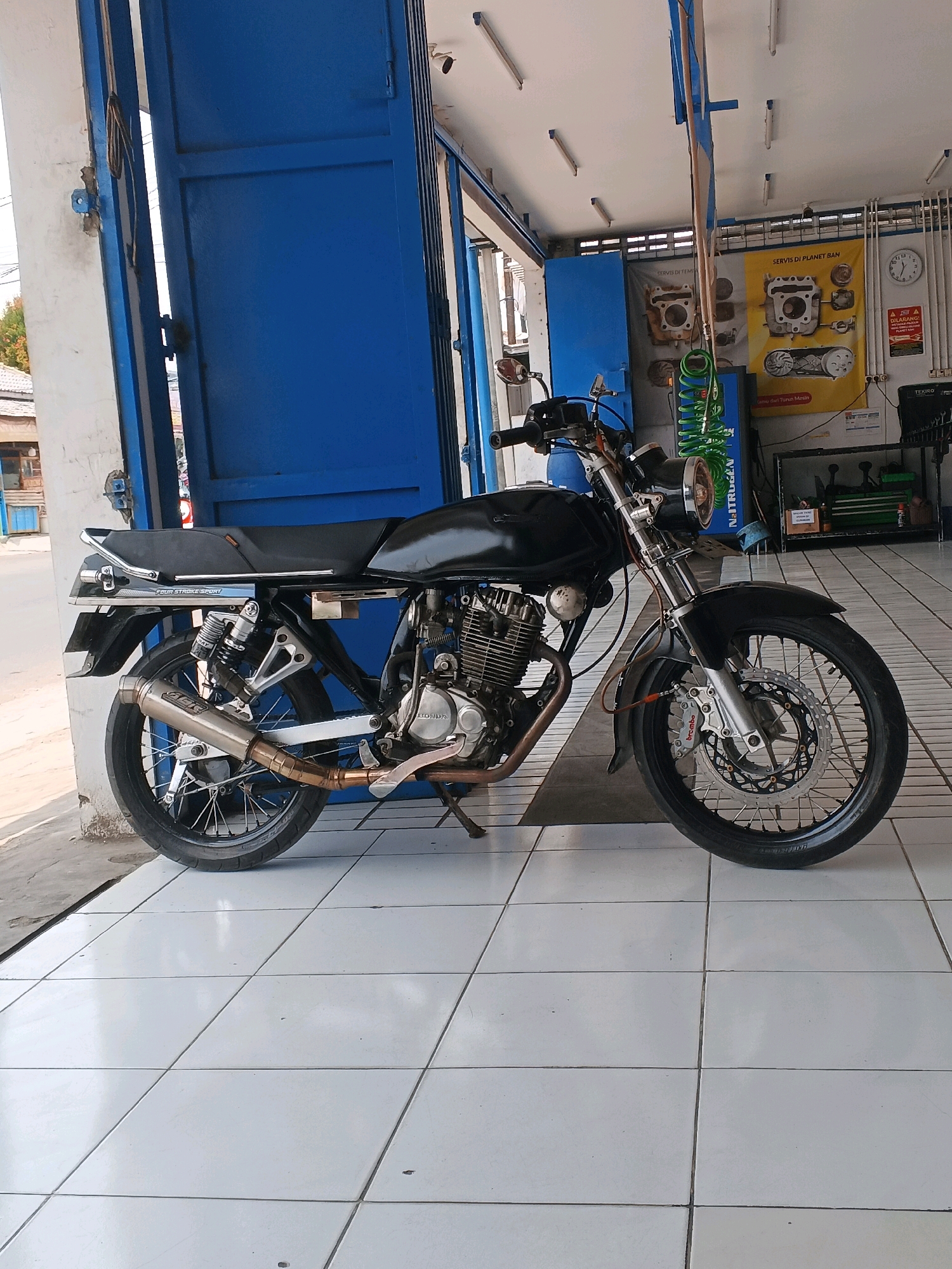 A black motorcycle is parked on a tiled floor in front of a blue garage door. The motorcycle is facing the right side of the frame. The garage door is open, revealing part of a workshop inside. The workshop has posters on the wall, tools on a shelf, and fluorescent lighting on the ceiling. The floor is made of white square tiles. The motorcycle has a chrome exhaust pipe and a black seat. The background shows a street with buildings and trees. The scene appears to be in a commercial area. The location is identified as Taman, Indonesia.