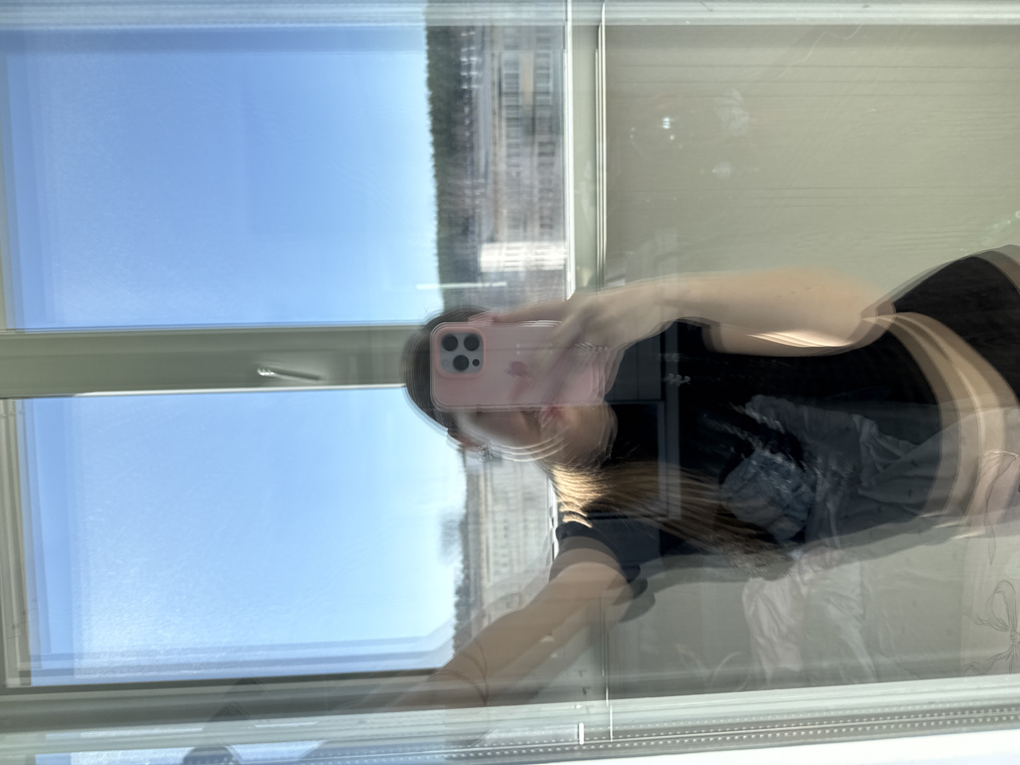A reflection in a window shows a person holding a pink iPhone, a view of a cityscape, and part of the interior. The background landscape, visible through the window, appears to be a skyline including a large building, trees, and a clear blue sky. The location is assumed to be Chaikovsky, Russia.