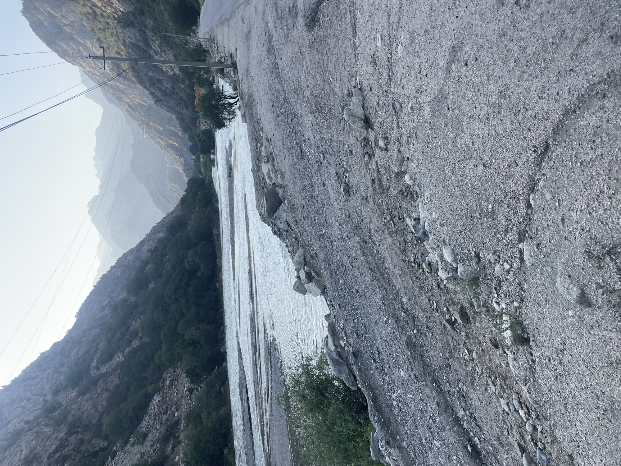 The image captures a view of the Varzob River in the city of Khodzha-Obigarm, Tajikistan. The river flows through a mountainous valley, with steep, rocky slopes covered in trees on either side. A paved road runs along the right bank of the river, with power lines overhead. The ground in the foreground is a mixture of gravel and sand, with some scattered rocks. The sky is clear and hazy, suggesting it may be early morning or late afternoon. The overall scene is one of natural beauty, with a sense of remoteness and tranquility. There are no people or animals visible in the image.
