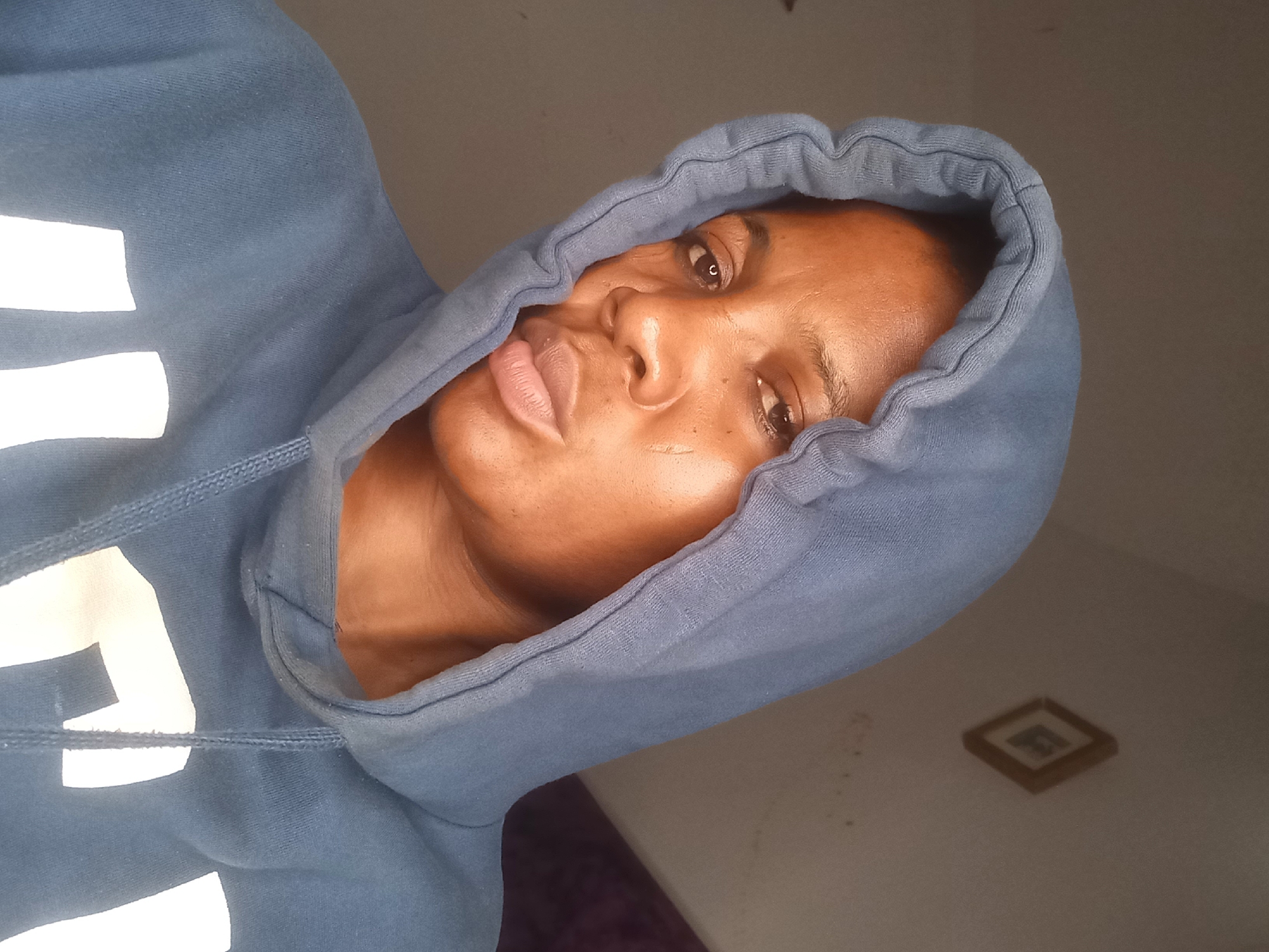 A portrait shows a Black woman wearing a blue hooded sweatshirt. She is positioned centrally in the frame, with her face angled slightly to her right and her head tilted. Her eyes are looking towards the camera. A portion of the wall behind her, a framed picture on the wall, and a hint of purple fabric are visible. The lighting in the image appears to be natural light coming from the left. The overall scene is indoors, and the backdrop suggests a domestic setting. There is no discernible urban context that would indicate a specific location within Jos, Nigeria.
