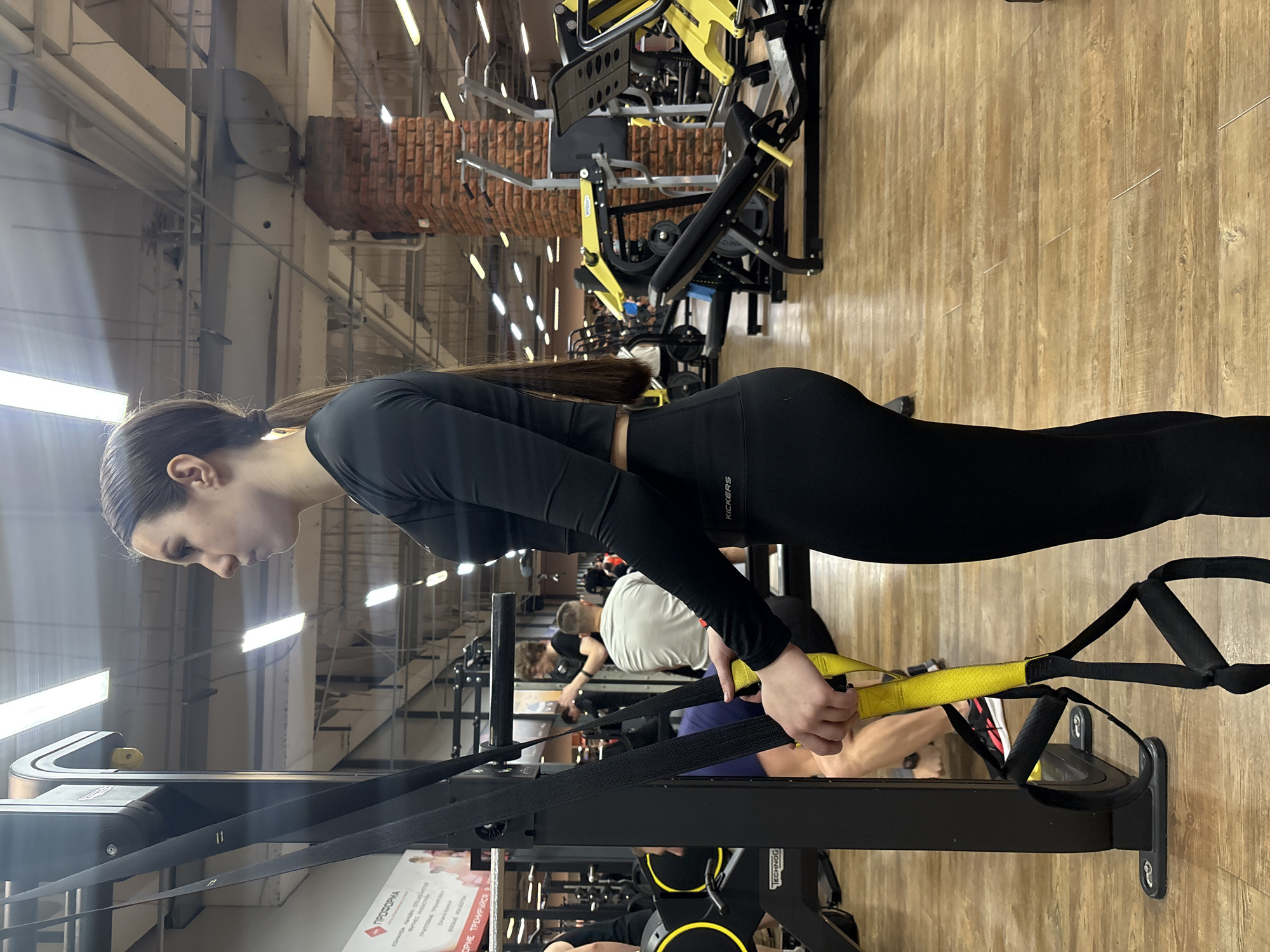 An image captures a young woman exercising in an indoor gym, oriented so she appears to be standing upright from the left of the frame. She is seen in profile, with dark hair tied back in a ponytail, wearing a black long-sleeved crop top and black leggings with "KICKERS" visible on the left leg. She holds the yellow and black handles of a suspension trainer (TRX-style), leaning back as if performing a pulling motion, with a focused expression.
The gym features a light-colored wooden floor and an industrial-style ceiling with exposed white beams, ductwork, and linear fluorescent lighting. Various black and yellow gym machines, such as leg presses and strength equipment, are visible in the blurred background. Several other individuals, primarily men, are also seen exercising or moving around. Decorative elements made of red brick are integrated into the gym's architecture. A white sign with red text, partially visible as "ПРОФОРМА" (PROFORMA) and other Russian words, is attached to the black frame of the suspension trainer setup. The scene depicts an active fitness environment in Chaikovsky, Russia.