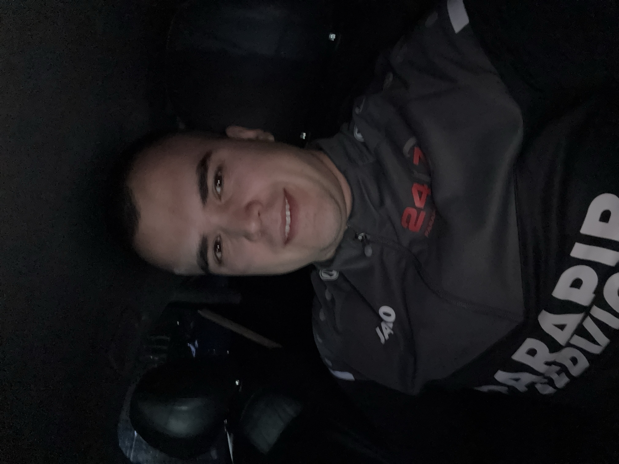 A selfie of a man wearing a grey jacket with "JAKO" and "24/7" logos, and a black t-shirt with "PARADIV" printed on it. The location is unknown, Unknown. The environment is dark, possibly indoors.