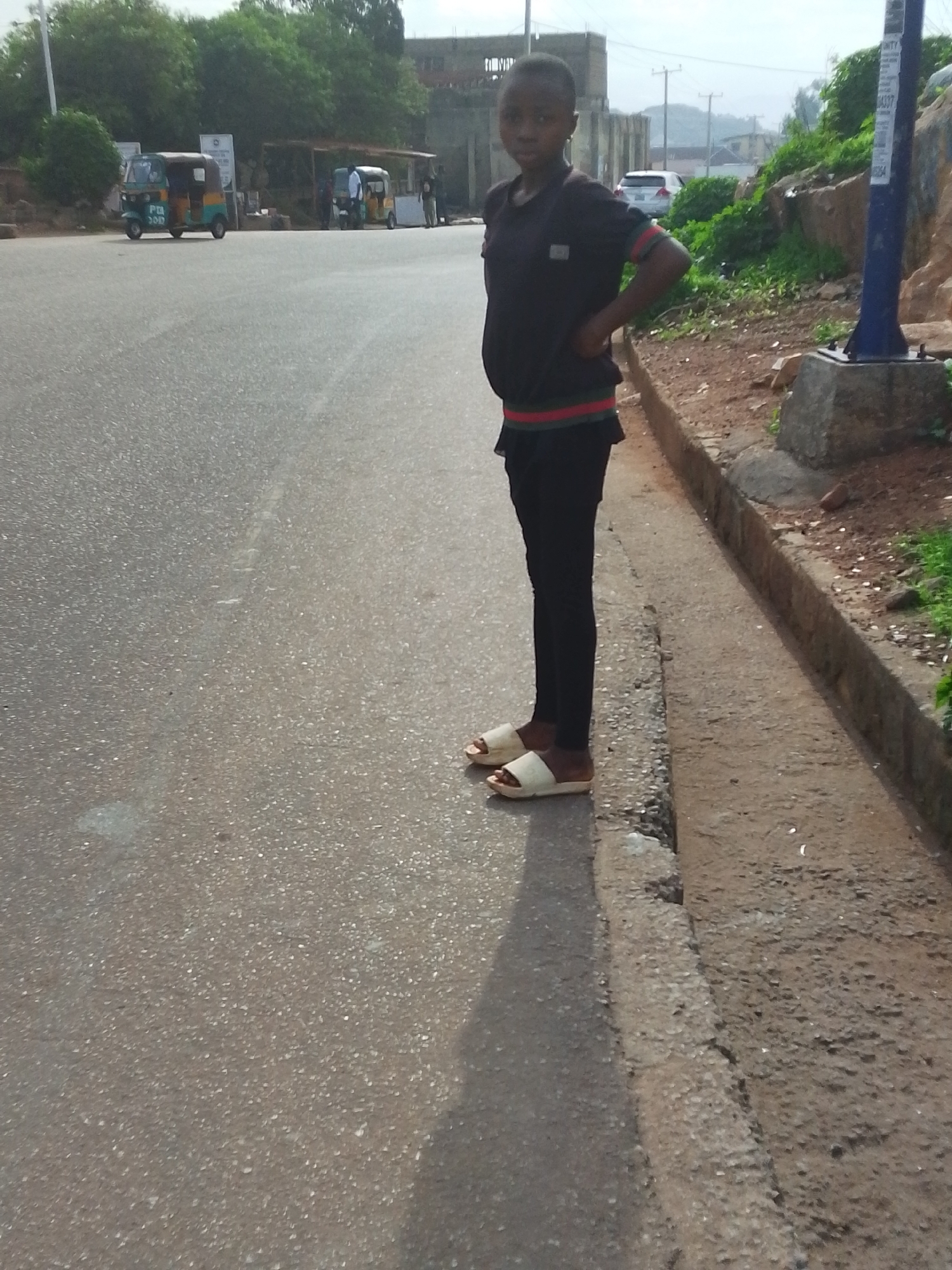 A young person stands on a paved roadside in Jos, Nigeria. They wear a dark top, black pants, and light-colored sandals. The road surface is asphalt and a concrete curb separates the road from a dirt area with greenery. In the background, there are several vehicles, buildings, and signs. A blue pole with notices is visible on the right of the frame.