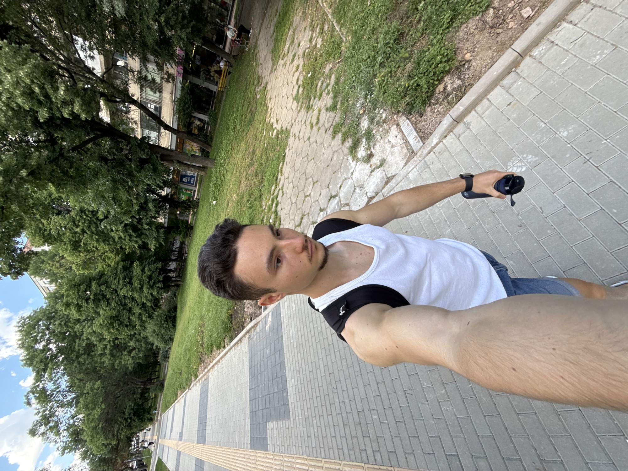 A selfie taken by a fair-skinned male with dark hair and a slight beard, wearing a white tank top with black straps and blue jean shorts, standing on a grey bricked walkway. He is holding a dark blue cylindrical object with a black band around his left wrist. The walkway is lined with green grass and trees on the left, leading to buildings in the background. On the right, the walkway is bordered by a strip of green grass and bushes, and beyond that, a stone-paved area. Based on the trees, architecture, and paving, the location is likely a public park or pedestrian area in Sofia, Bulgaria.