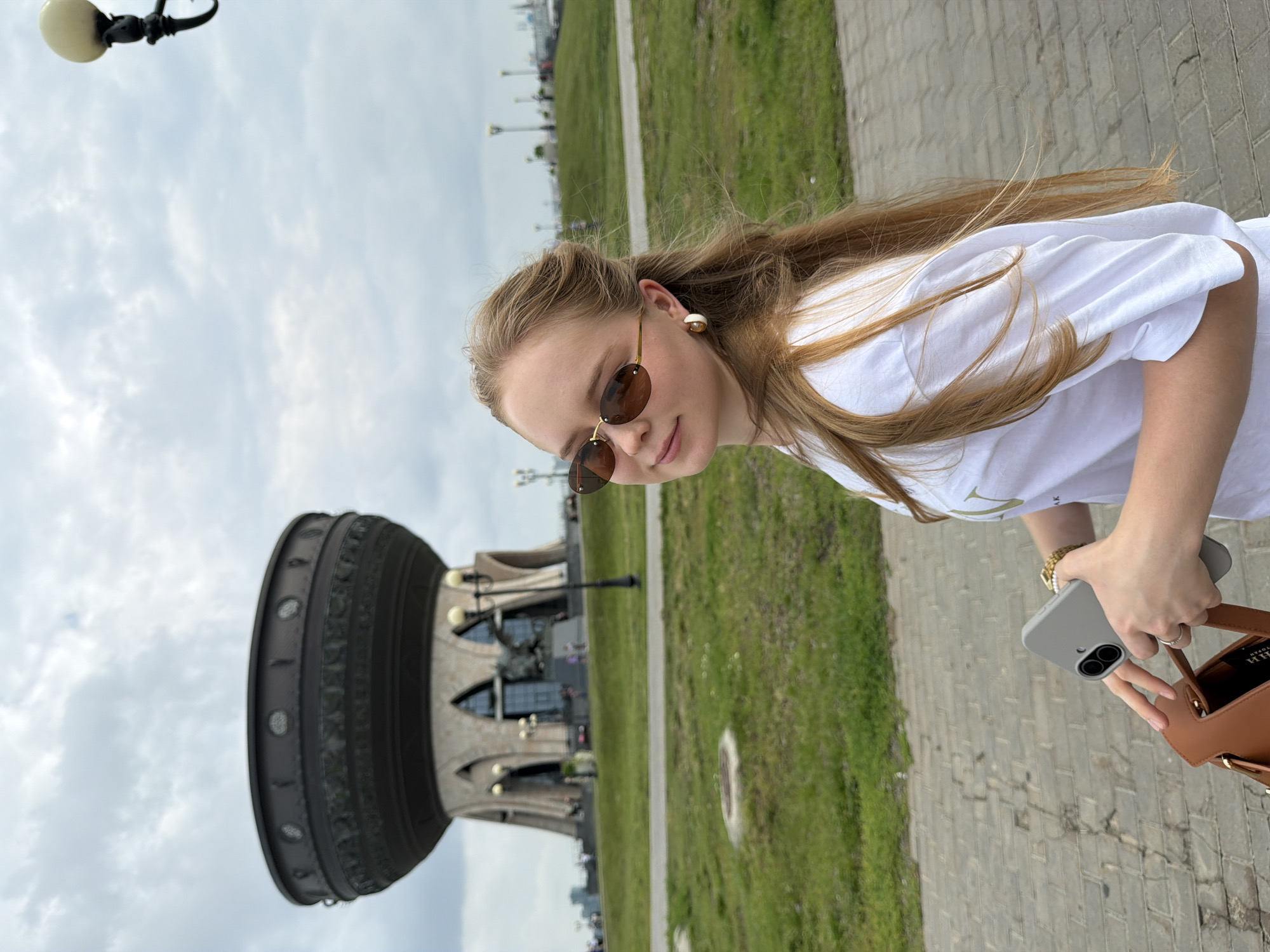 A young woman with long blonde hair is standing on a paved walkway in front of a large, distinctive building. She is wearing sunglasses, a white t-shirt, and earrings. She is holding a smartphone in her left hand and a brown bag in her right hand. The building behind her is shaped like a large cauldron or pot, with ornate carvings and a light-colored base. It is situated on a grassy hill, with a cloudy sky overhead. This is the Family Center "Kazan" in Kazan, Russia. The surrounding area appears to be a park or public space with lampposts and other greenery.