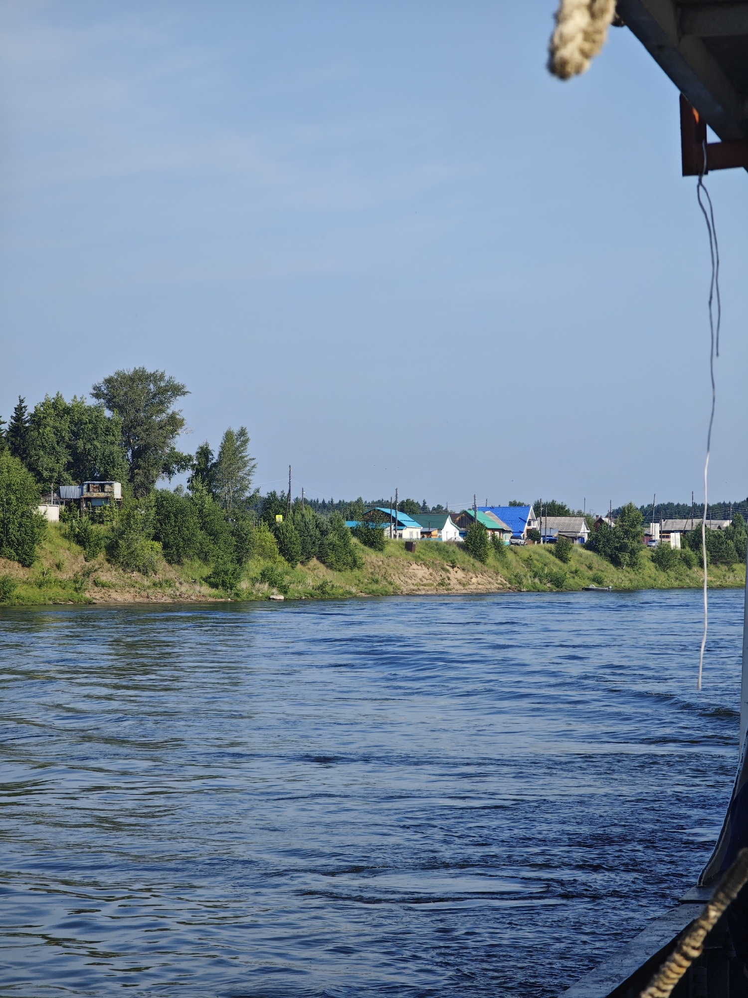 The image shows a view of the river in Galanino, Russia, from a boat. The water is blue and choppy, with ripples and waves. On the far bank, there is a line of trees and houses, with green roofs and white walls. The houses appear to be in a village, with power lines running between them. The sky is a clear blue, with a few wispy clouds. In the foreground, part of the boat is visible, with a thick rope tied to it. The overall impression is one of a peaceful, rural scene.