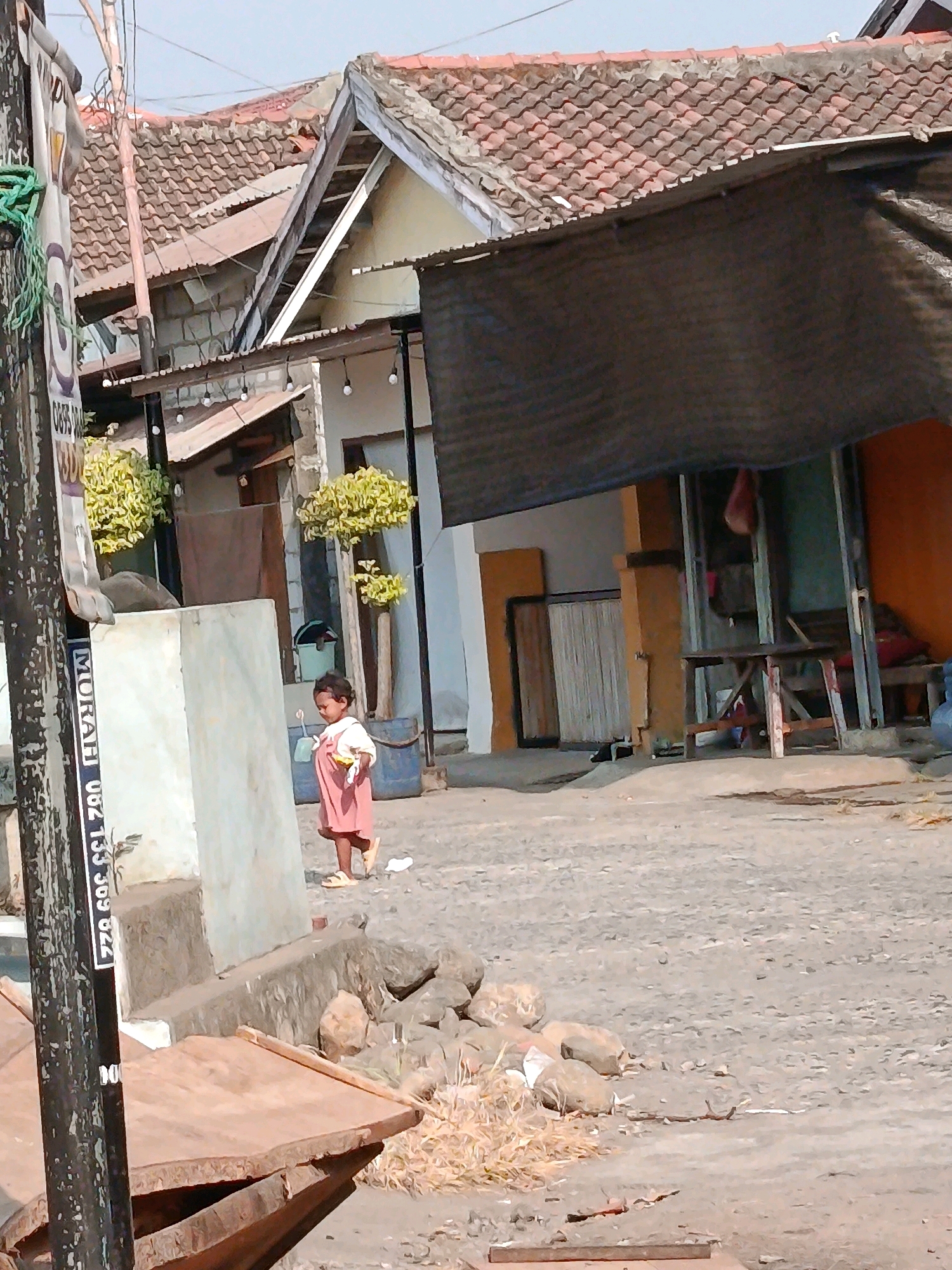 The image captures a street scene in Jobokuto, Indonesia, under a clear, bright sky. In the foreground, a small child, appearing to be a girl with dark hair, is walking. She is wearing a light pink dress or romper and light-colored sandals. The child is holding a yellow object in her right hand. The ground is paved with small stones and gravel. To the left of the child, a vertical black pole with white lettering is visible, with green rope hanging from the top. Behind the child, a light-colored concrete structure or wall is present. Further back, several buildings with tiled roofs line the street. One building has a dark, opaque awning stretched over its front. The buildings appear to be a mix of residential and commercial structures. Small trees with yellow-green foliage are planted in front of some of the buildings. The overall environment suggests a typical street in a residential or mixed-use area of an Indonesian town.