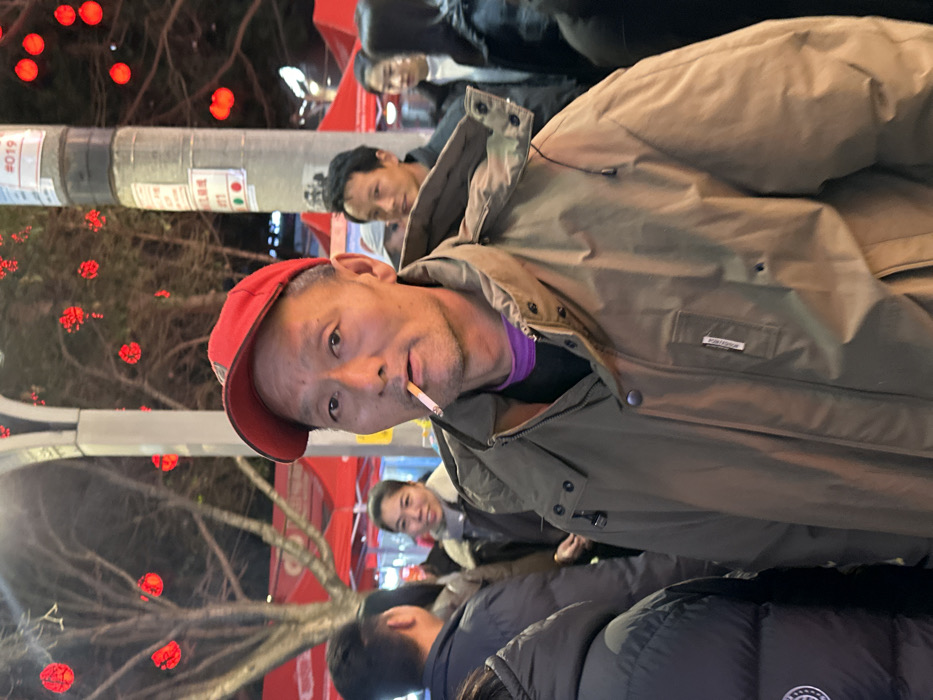 This image depicts an outdoor night scene in the city of Jiangjin, China, portraying a busy public area. The central figure is an adult East Asian male, positioned in the foreground and looking directly at the camera. He wears a red baseball cap, a purple undershirt, and a tan winter jacket featuring a white brand tag on its left chest pocket. An unlit cigarette is held between his lips. In the blurred mid-ground and background, several other individuals are visible, including a smiling woman to the left of the main subject and a man to his right. The environment is illuminated by artificial lighting, highlighting numerous red spherical lanterns or decorative lights suspended from trees and overhead structures, alongside red market awnings or tents. A decorative, light-colored street lamp column is visible on the left, and a dark utility pole on the right bears multiple affixed signs, including one marked "019" and others with red and green circular indicators. The scene suggests a bustling evening market or public gathering.