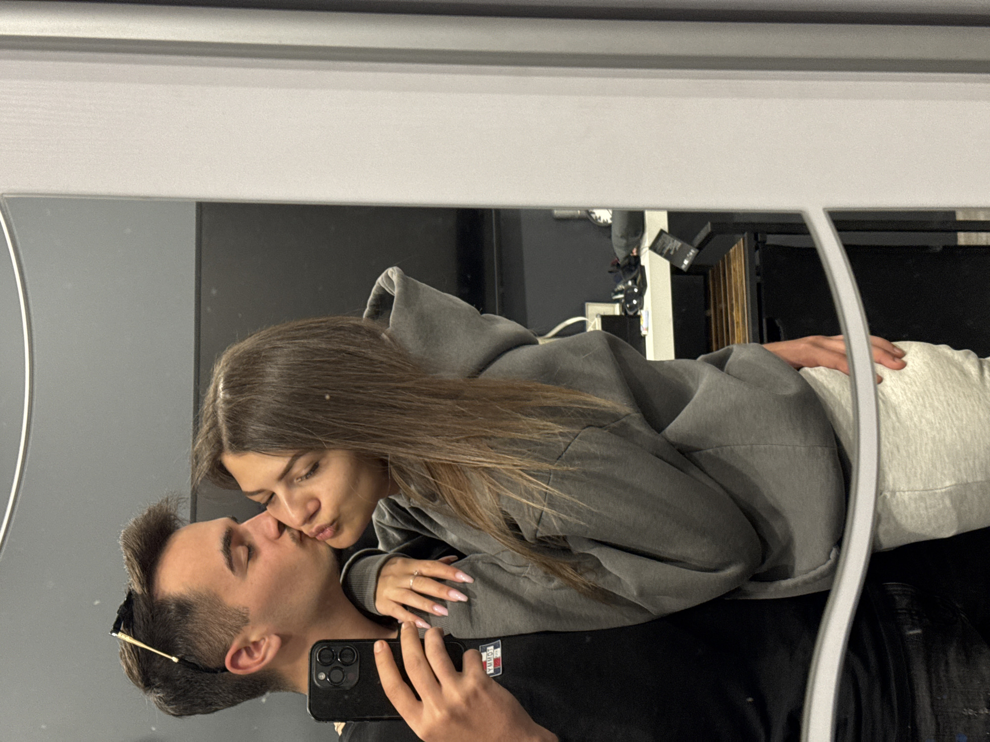 The image, taken in Sofia, Bulgaria, captures a mirror selfie of a young couple displaying affection. The photograph is oriented horizontally, showing the subjects in a reclining or leaning posture.
The woman, with long brown hair, wears a grey hooded sweatshirt and light grey sweatpants. She is leaning over the man, puckering her lips in a kissing gesture directed towards his cheek, with her eyes closed or partially closed. Her left hand rests gently on the man's chest, adorned with a ring on her ring finger and light pink manicured nails.
The man, with short dark hair and sunglasses tucked into the collar of his black t-shirt, is holding a black smartphone with multiple camera lenses (likely an iPhone) to capture their reflection. A small white logo, resembling a Tommy Hilfiger emblem, is visible on his t-shirt. His eyes are also closed or partially closed.
The setting appears to be indoors, viewed through a large mirror with a prominent white and grey frame along the top edge of the image. A curved, vertical light strip is visible on the right side of the mirror's reflection. The background reflected in the mirror beyond the couple consists of dark surfaces, possibly shelves or furniture, with some indistinct objects visible in the upper right. The overall atmosphere conveyed is intimate and affectionate. No specific time of day is discernible.