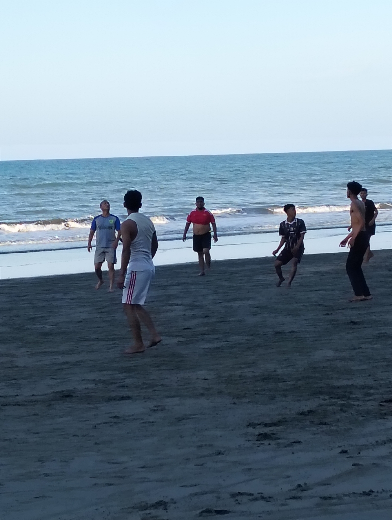 A group of six young men are playing soccer on a sandy beach next to a calm ocean under a clear, pale blue sky. The man in the foreground on the right is wearing black pants and is bare-chested. To his left and slightly behind him is a young man in a black and white soccer jersey and black shorts, in mid-action as if kicking a ball. Further to the left, another man in a white tank top and white shorts with red stripes is looking up. He is the tallest person visible and appears to be in motion. On the far left, a man in a grey t-shirt and grey shorts is looking towards the sky. In the middle distance, a man in a red t-shirt and black shorts is also looking up. The ocean is a deep blue with small waves lapping at the shore. The sand is a dark grey color, showing footprints and slight indentations. The lighting suggests it is either early morning or late afternoon.