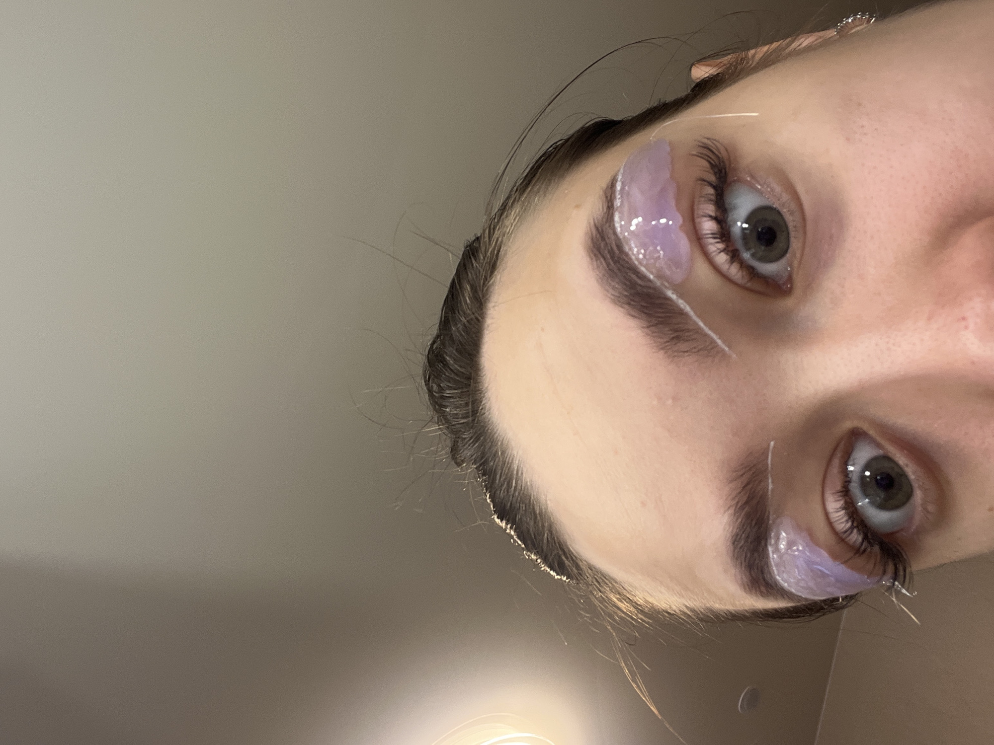 A low-angle, close-up, indoor photograph shows the upper portion of a person's face, from the eyebrows down, with a pale, neutral background that is out of focus. The person has dark brown hair pulled back, and their eyebrows appear to be treated with a light purple substance and white tape. Long, dark eyelashes are visible on both eyes. The person's eyes are open and looking directly at the camera, with green irises and dark pupils. The skin is fair and appears smooth. The lighting is soft and coming from the left side of the image, casting a subtle glow on the hair and forehead. The image does not contain any information that would indicate its location in the city of Chaikovsky, Russia.