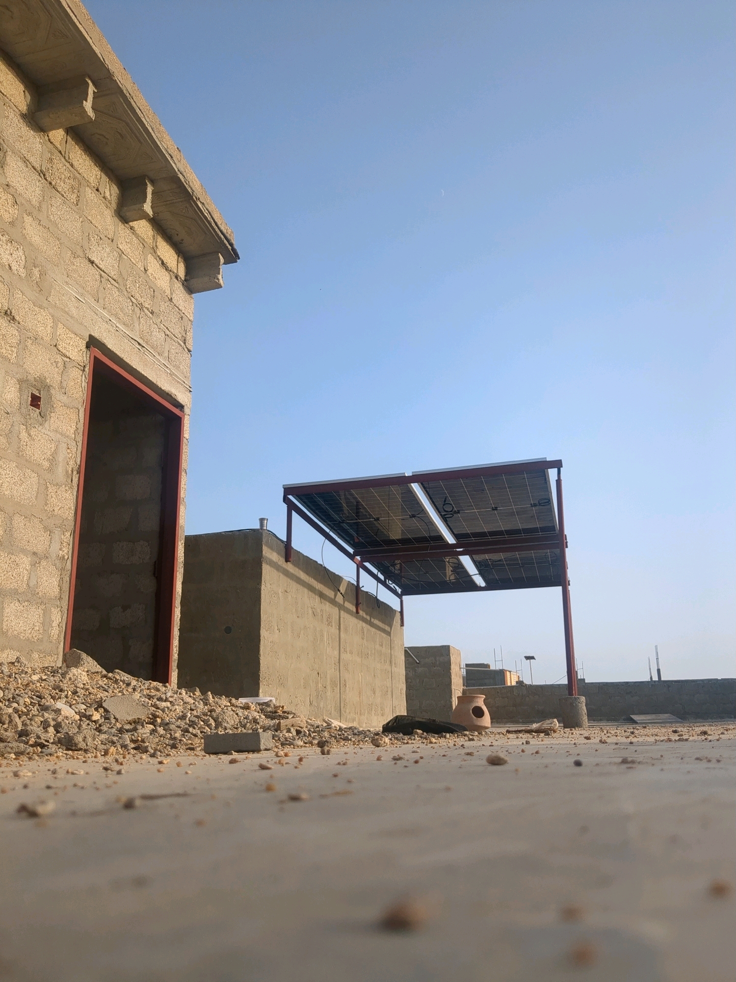 The image depicts an outdoor scene on a rooftop, likely in Karachi Division, Pakistan, characterized by a clear blue sky. In the foreground, a concrete surface is littered with small stones and debris, suggesting an unfinished construction or a storage area. To the left, a partially constructed building made of light-colored bricks is visible, with an open doorway framed by red metal. Above this building, decorative concrete elements adorn the roof's edge. Further into the scene, a concrete structure with a wall is prominent. Mounted on a red metal frame above this wall are solar panels, indicating a focus on renewable energy. In the background, more of the rooftop landscape unfolds, featuring various construction elements, including concrete walls and what appears to be a clay pot with a circular opening. The overall impression is one of an urban environment in a state of development or ongoing activity, with a mix of traditional building materials and modern technology.