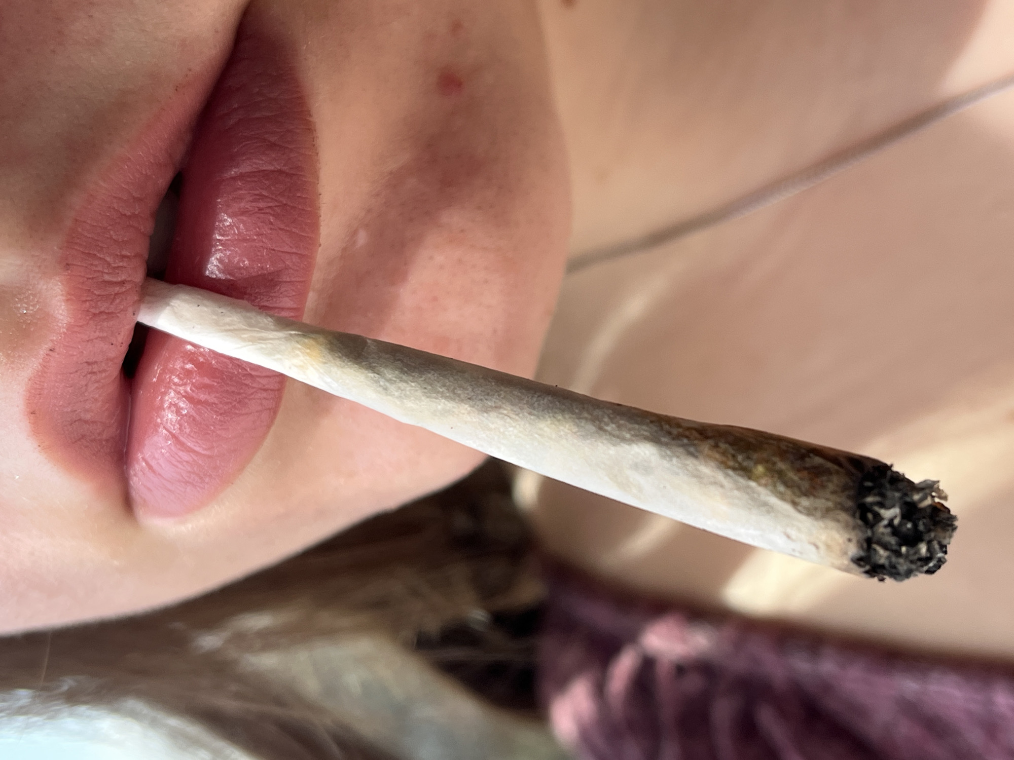 A close-up image shows a person's face with a lit joint held in their mouth. The subject has full, pink lips and fair skin. Part of their neck and shoulder are visible, with a hint of purple clothing. The joint is partially smoked, with burnt residue at the end. The location is unknown and there is no evidence to suggest this image was taken in Varna, Bulgaria.