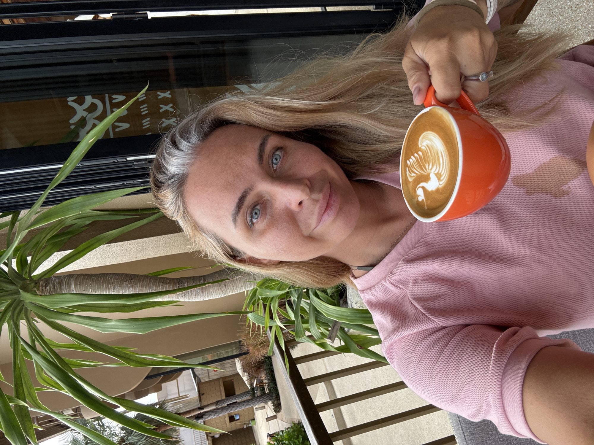 A woman with blonde hair and blue eyes, wearing a pink sweatshirt, holds an orange mug of latte art featuring a swan. She is seated outdoors, with a palm tree and a building visible in the background. The location appears to be a cafe in Ko Samui District, Thailand, judging by the tropical foliage and architectural style. The woman is looking at the camera and smiling slightly.