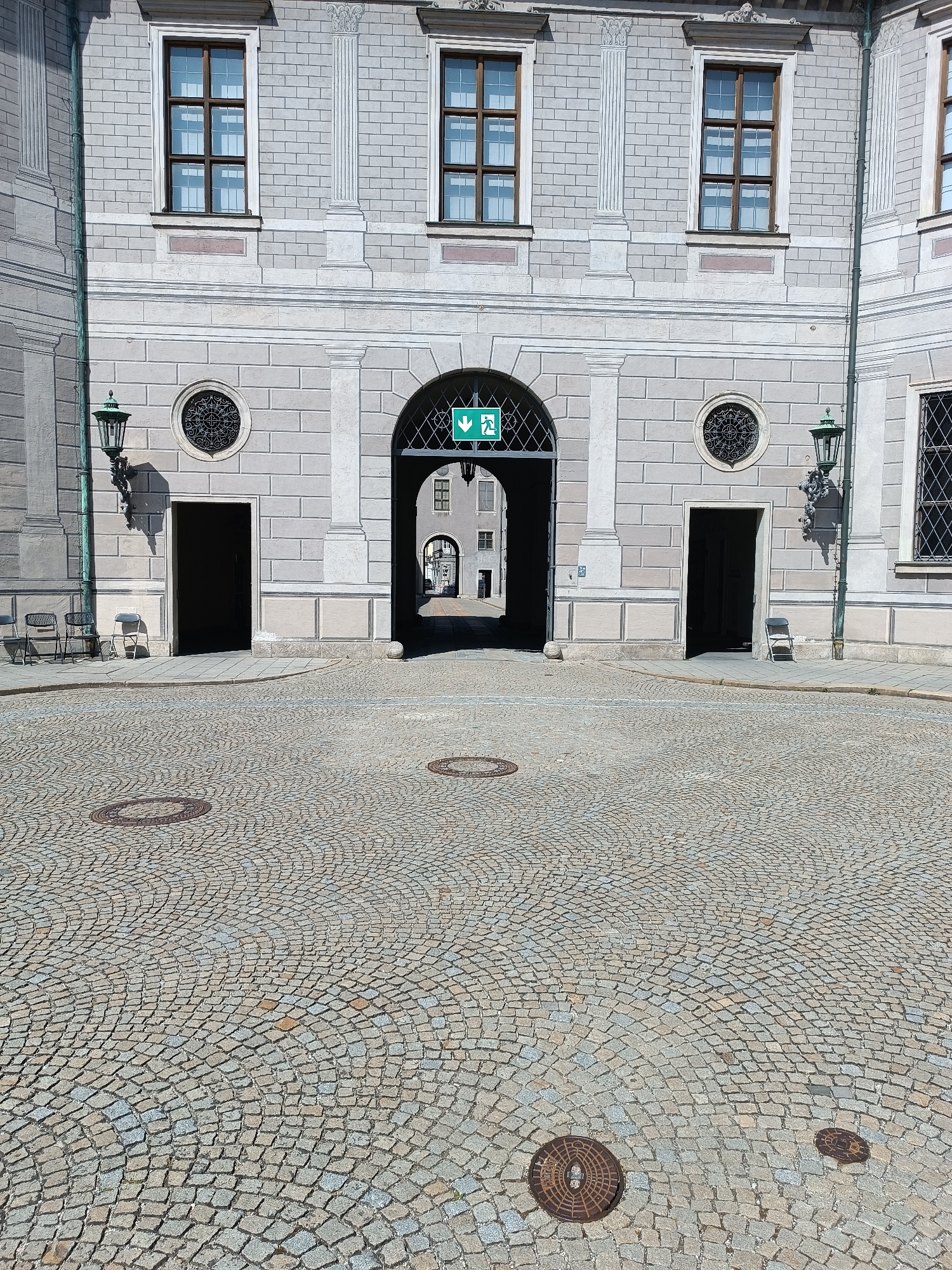 The image shows a cobblestone courtyard in front of a light gray stone building in Munich, Germany. The building features an arched entryway in the center, flanked by two dark doorways and two round decorative windows. Above the doorways, there are rectangular windows with dark frames. A green emergency exit sign with an arrow pointing downwards is visible above the central arch.
In the courtyard, there are two manhole covers visible on the cobblestones. To the left of the central archway, near a green lamppost, are two chairs. To the right of the central archway, near another green lamppost, is a single chair positioned in front of the dark doorway. The courtyard appears to be empty of people, aside from the potential implied presence of occupants within the building or those who have used the chairs. The lighting suggests it is daytime with the sun casting shadows.