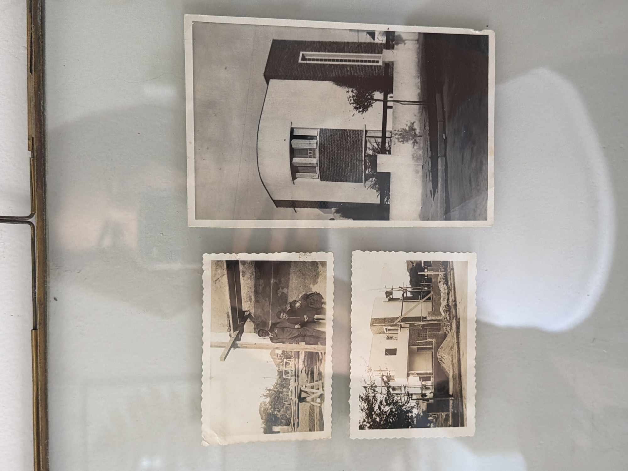 The image shows three black and white photographs, arranged in a glass frame with a metal border. The largest photograph is oriented horizontally and depicts a building with a curved facade and rectangular windows, likely a residence. Two smaller photographs, both with scalloped edges, are positioned below the larger one. The photograph on the left shows two adults and a child standing in what appears to be a garden or outdoor space, likely in a family portrait. The photograph on the right depicts the construction of the same building, with scaffolding visible. Vicente López, Argentina, is presumed as the location based on prior knowledge or implicit information.