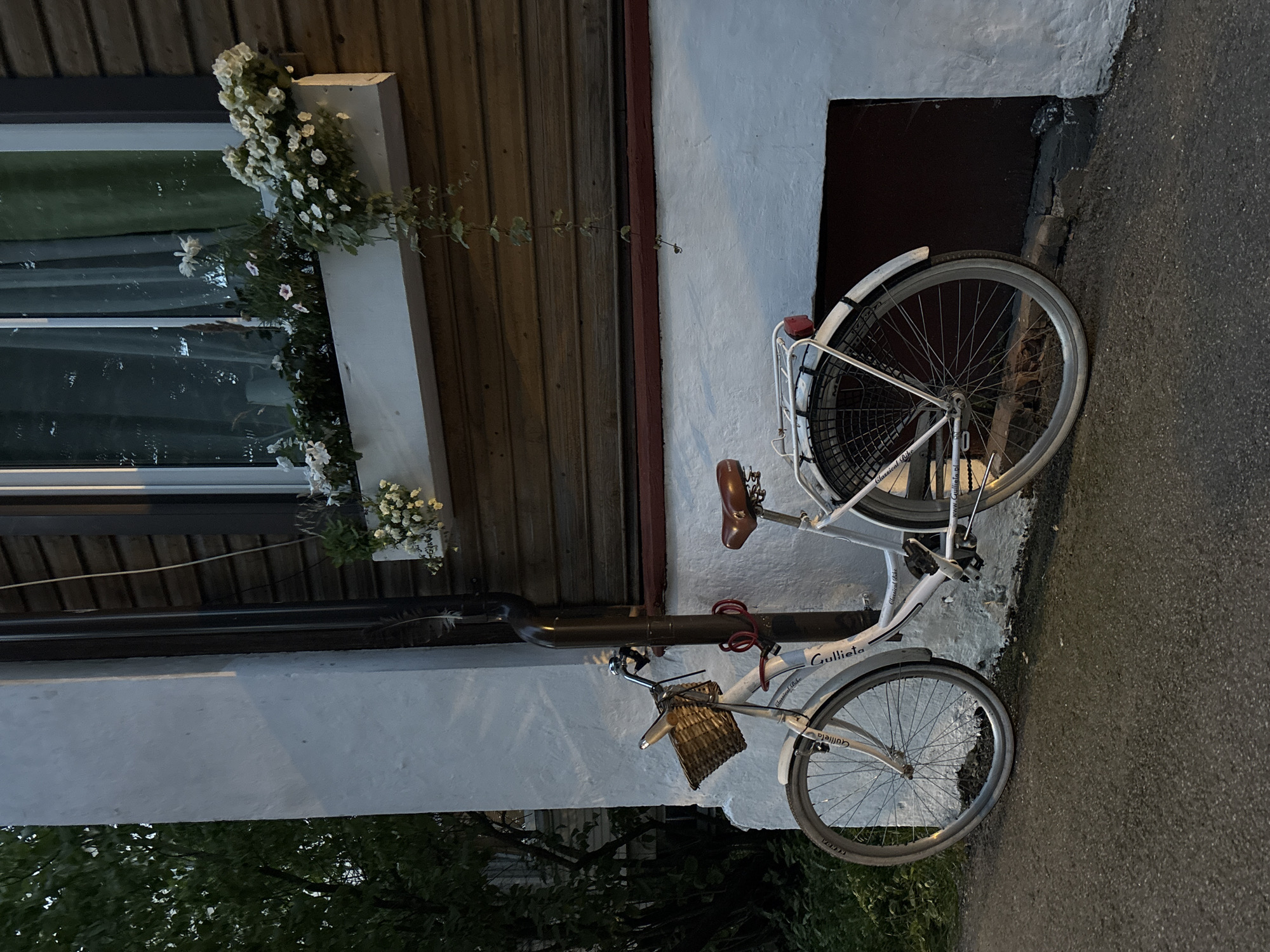 A white bicycle with a brown saddle and a wicker basket is parked leaning against a textured white wall. The bicycle has a red U-lock attached to its frame and a red taillight. Above the bicycle, a window box filled with white flowers is mounted on a dark wooden wall. A dark downspout runs vertically along the white wall, next to the bicycle. The scene appears to be on a street in the city of Tallinn, Estonia. The lighting suggests it is either early morning or late evening.