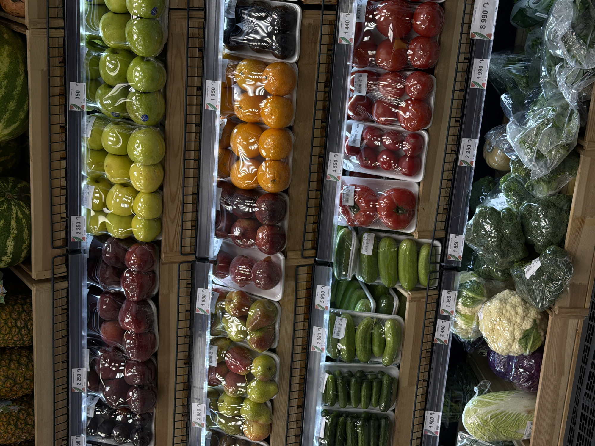The image shows an assortment of fruits and vegetables displayed in a refrigerated section of a supermarket, likely located in Yerevan, Armenia. The produce is arranged in neat rows with individual items or small groups packaged in clear plastic wrap and placed in white styrofoam trays.
The leftmost section contains watermelons and pineapples. Next to it, several rows of green apples, red plums, oranges, and pears are stacked vertically. Moving right, a row of red tomatoes is visible, followed by rows of green cucumbers and a final section featuring various types of lettuce, broccoli, and cauliflower. Each item appears to have a price tag attached, indicating the cost of each packaged portion. The display shelves are metallic, with a lattice structure.
