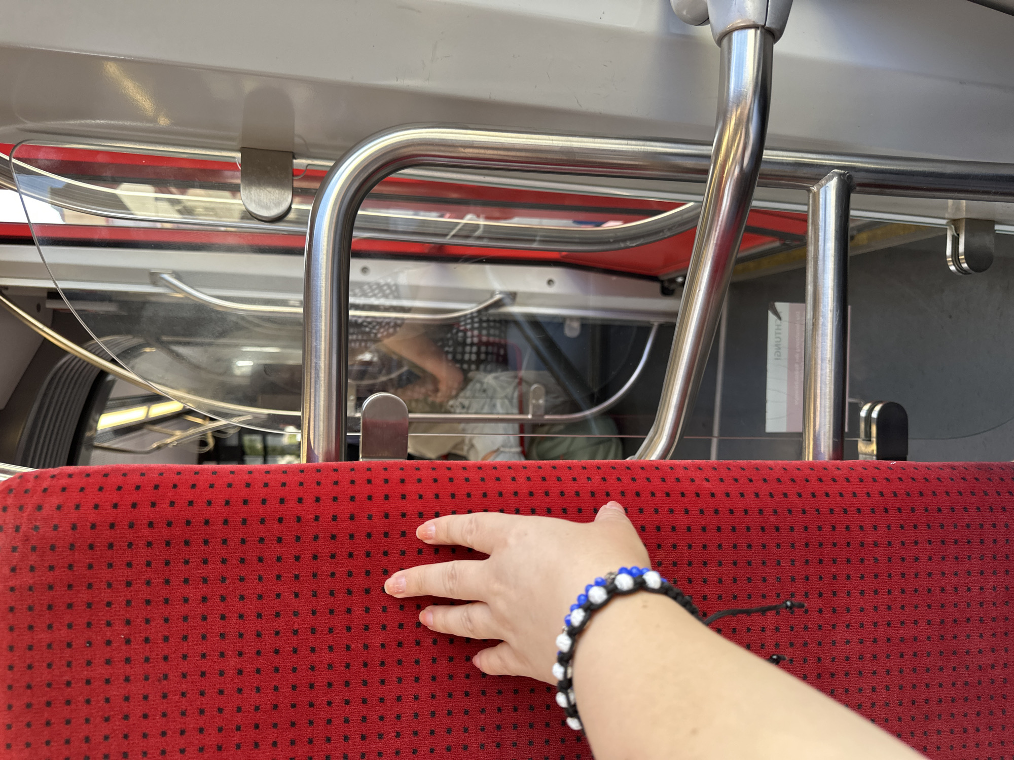 An image taken from inside public transportation, likely a tram or train in Krefeld, Germany, shows a person's left hand resting on a red seat with a black polka dot pattern. The hand is adorned with a bracelet featuring black, white, and blue beads. Behind the hand, a stainless steel handrail and a transparent partition are visible. In the background, reflections in the glass suggest the presence of other passengers and the exterior environment. The scene appears to be in motion, with blurred elements indicating movement.